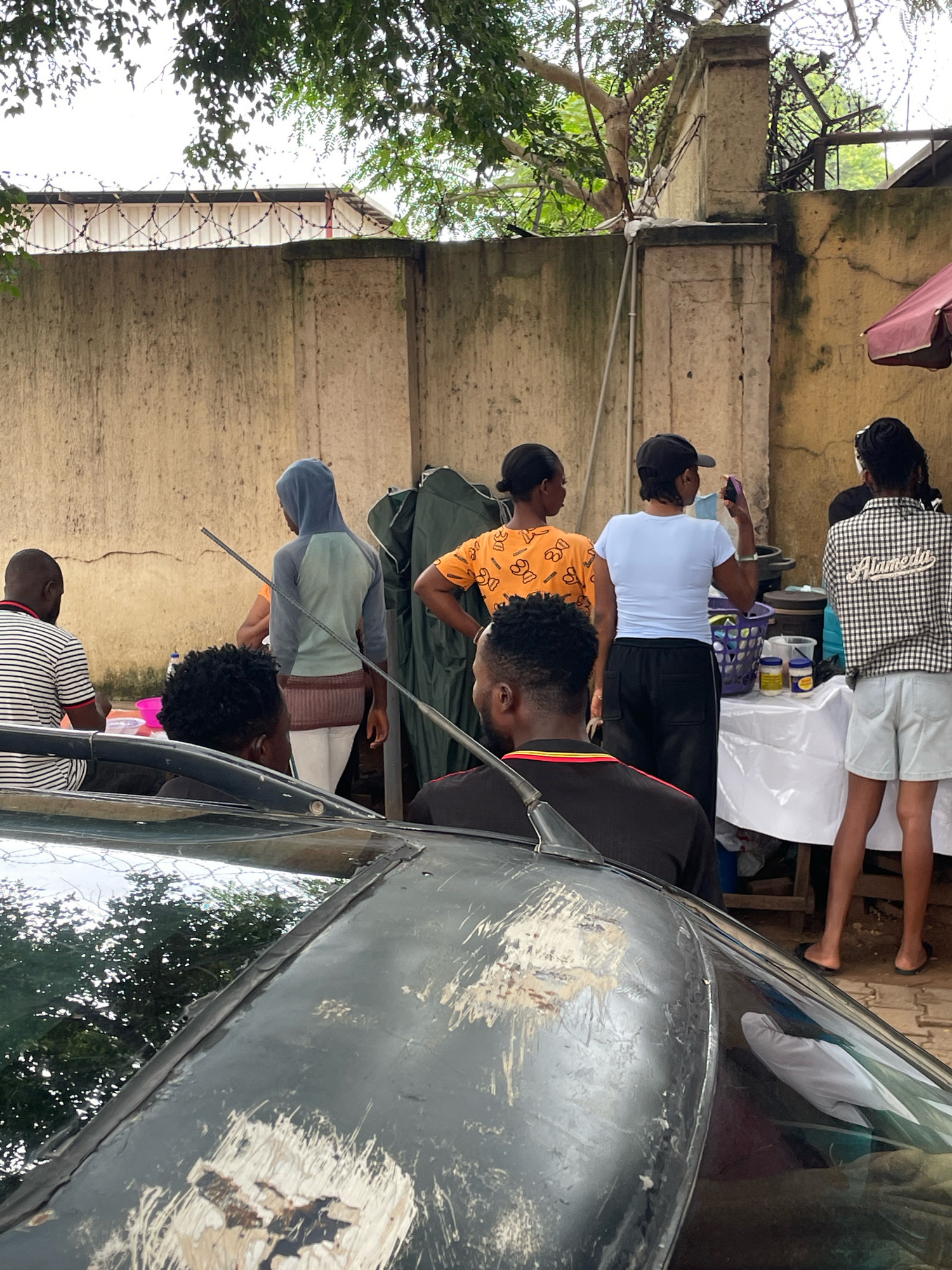 An outdoor scene depicts a group of people gathered near a wall with barbed wire on top. In the foreground, the hood and windshield of a dark-colored car are visible, showing signs of wear and tear. Behind the car, several individuals are interacting. One person, wearing a striped shirt, is seated and looking to their left. Another, with curly hair, is partially visible, facing away from the camera. A person in a blue hooded sweatshirt and white pants stands near a long metal rod. To their right, a woman in an orange shirt with a pattern is facing away. Further to the right, a woman in a light blue t-shirt and black pants holds a dark object, possibly a phone, near her face. Another woman, in a checkered shirt and light-colored shorts, stands to the far right. A table covered with a white cloth is set up, displaying various items including baskets and containers. The background features a textured, light-colored wall and green foliage overhead. The scene appears to be in a bustling area, possibly a street or market, in Abuja, Nigeria, given the distinctive architecture and surrounding environment.