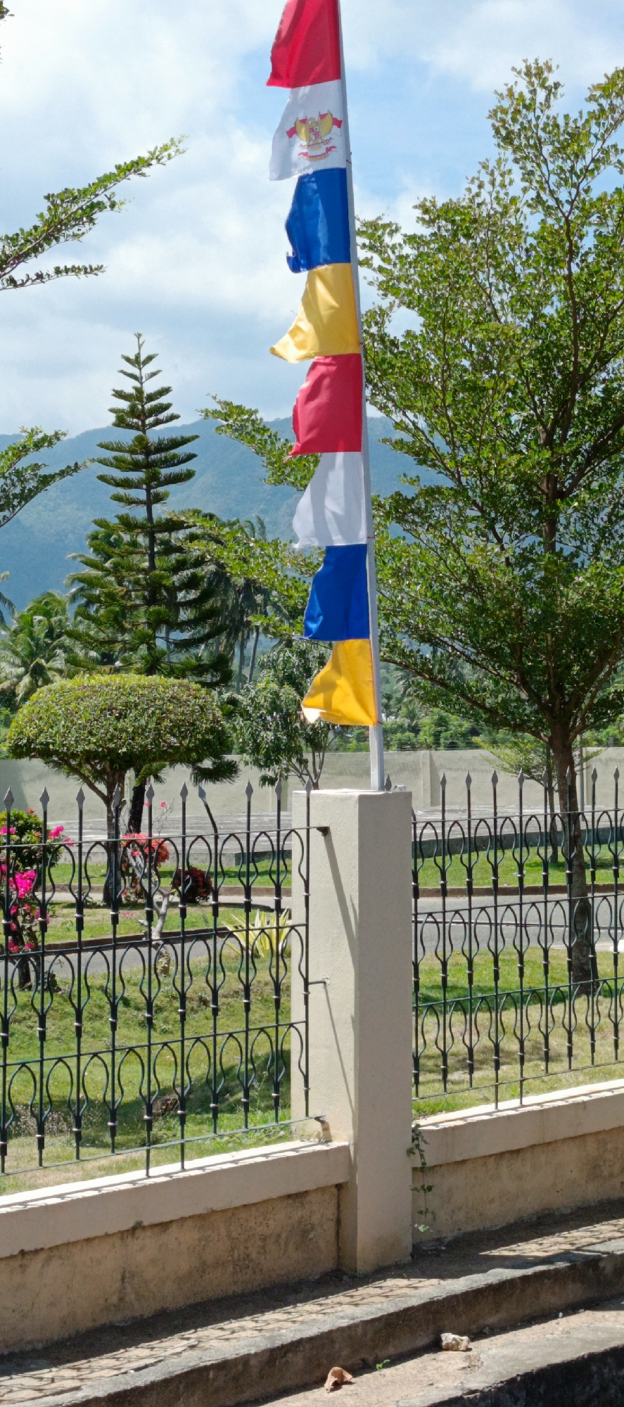 In Lampisang, Indonesia, a flagpole displays a sequence of colorful flags against a backdrop of lush greenery and distant mountains. The flags, from top to bottom, are red, white with a crest, blue, yellow, and red. A decorative iron fence runs horizontally across the mid-ground, separating a grassy area from a concrete pathway. Several trees, including a tall, conical evergreen and a broader, leafy tree, are visible. The sky is partly cloudy. The scene is tranquil and bathed in natural light.