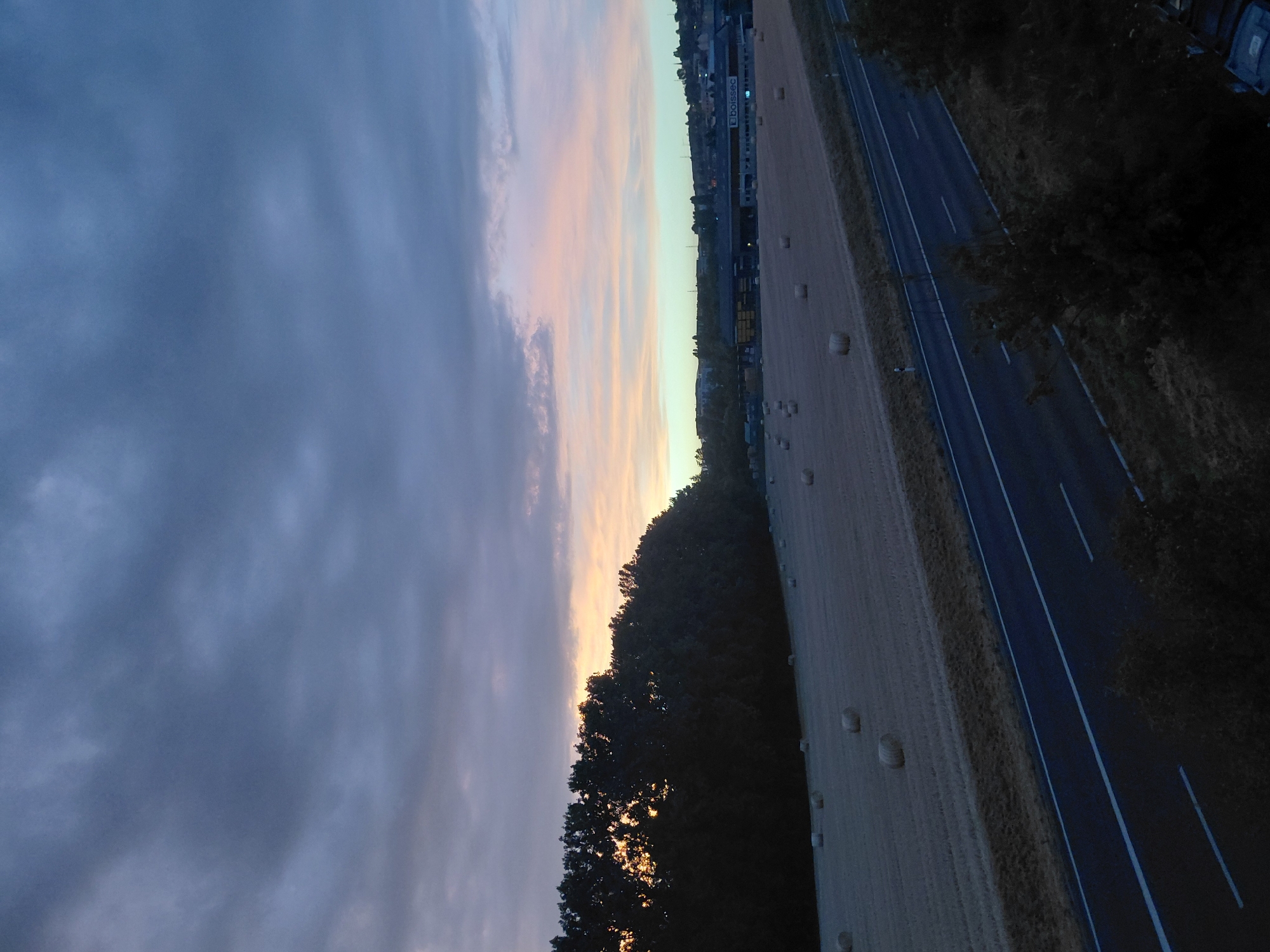 The image depicts a rural landscape near Ecublens, Switzerland, at dusk. In the foreground, a two-lane road with white markings runs diagonally from the bottom left to the bottom right. Adjacent to the road, on the left, is a field with numerous round bales of hay scattered across it. A dark line of trees runs along the far edge of the field, separating it from the horizon.
In the mid-ground, a low-rise industrial or commercial building complex is visible. It features a light-colored facade with dark windows and signage that appears to read "boissec". Beyond the buildings, a slightly undulating hillside is covered with scattered houses and more trees, forming a distant treeline against the sky.
The sky is predominantly filled with dramatic, dark gray clouds, especially in the upper portion of the image. Towards the horizon, there is a soft gradient of light, with hues of pale orange, pink, and a pale greenish-blue suggesting the last light of sunset or the first light of dawn. The overall impression is of a twilight scene over a cultivated agricultural area bordering a developed commercial zone. There are no people visible in the image.