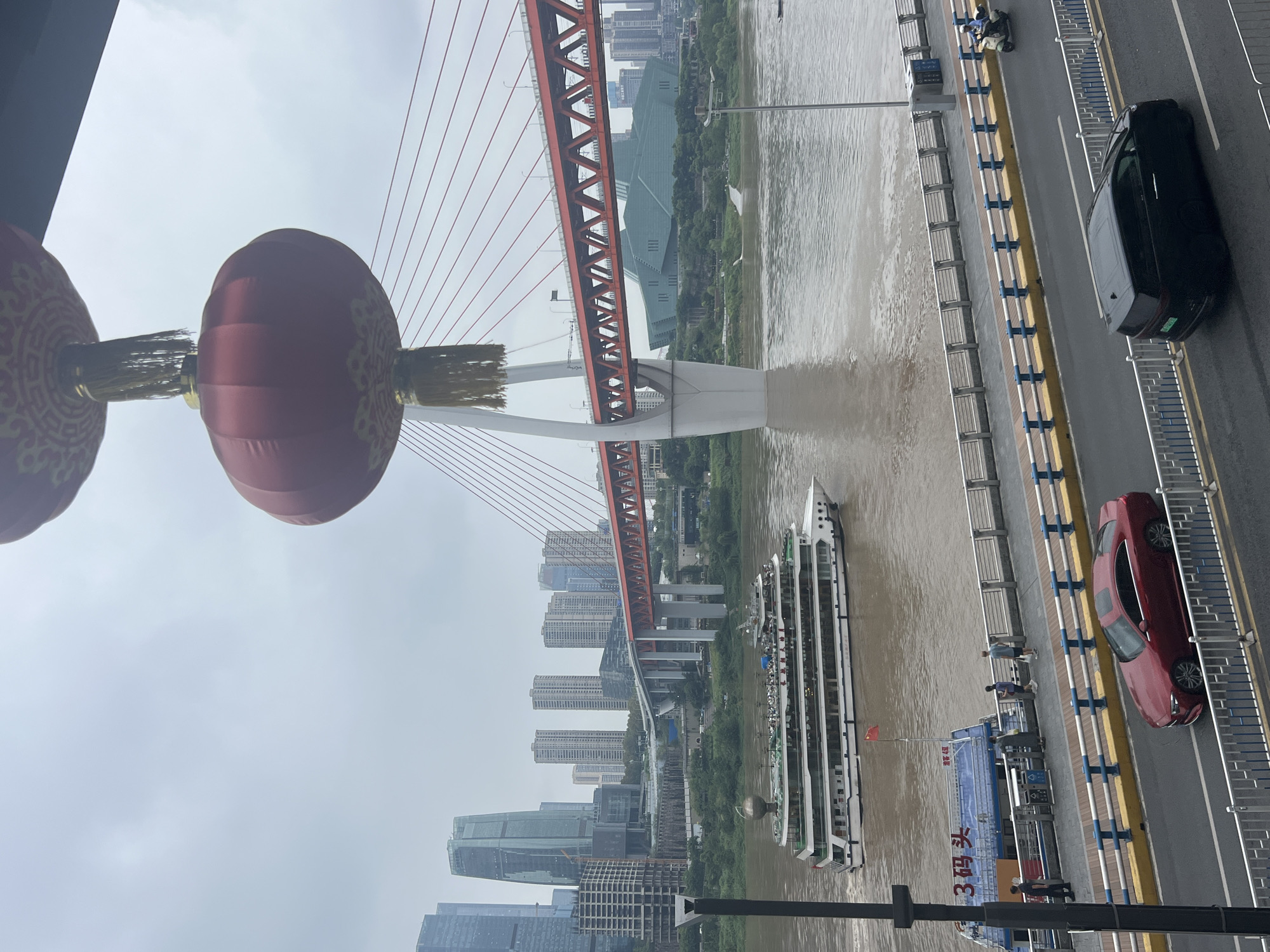 This is a medium shot of the Yangtze River in Chongqing, China, taken from an elevated perspective. In the foreground, a red sedan and a black van drive on a road, separated by a white fence. Behind the road is a pedestrian walkway with people strolling. A large tour boat is on the river, and a smaller ferryboat is docked nearby. In the background, a large red suspension bridge crosses the river, with skyscrapers and buildings of varying heights visible on either side. Two large red lanterns hang from the top of the frame. The sky is overcast, and the water is a murky brown. The overall impression is of a busy urban riverfront scene.
