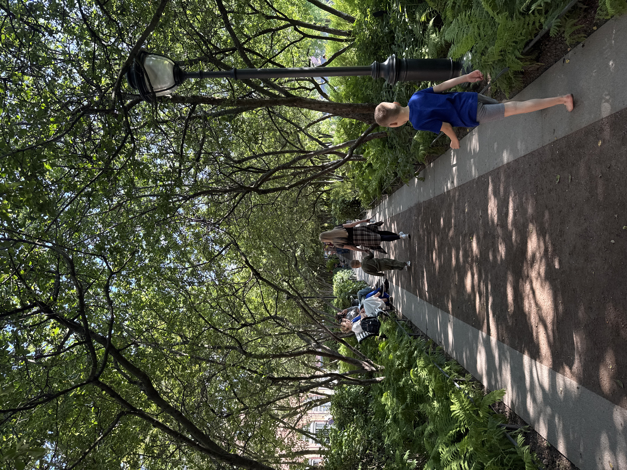 The image depicts a sun-dappled park scene, likely in an urban setting given the presence of a lamppost and buildings visible in the background. A paved pathway, bordered by lush green ferns and trees with dense foliage, runs through the center of the frame. Several people are present in the park. In the foreground, a young boy wearing a blue t-shirt and gray shorts walks barefoot on the path, with his back to the camera. Further down the path, a woman with long blonde hair, wearing a plaid shirt and black leggings, walks ahead of a young boy in a green jumpsuit. To the left, a group of people are seated on a park bench, enjoying the shade of the trees. The overall atmosphere is peaceful and serene, with sunlight filtering through the leaves, creating a play of light and shadow on the ground. The location is described as Unknown, Unknown.