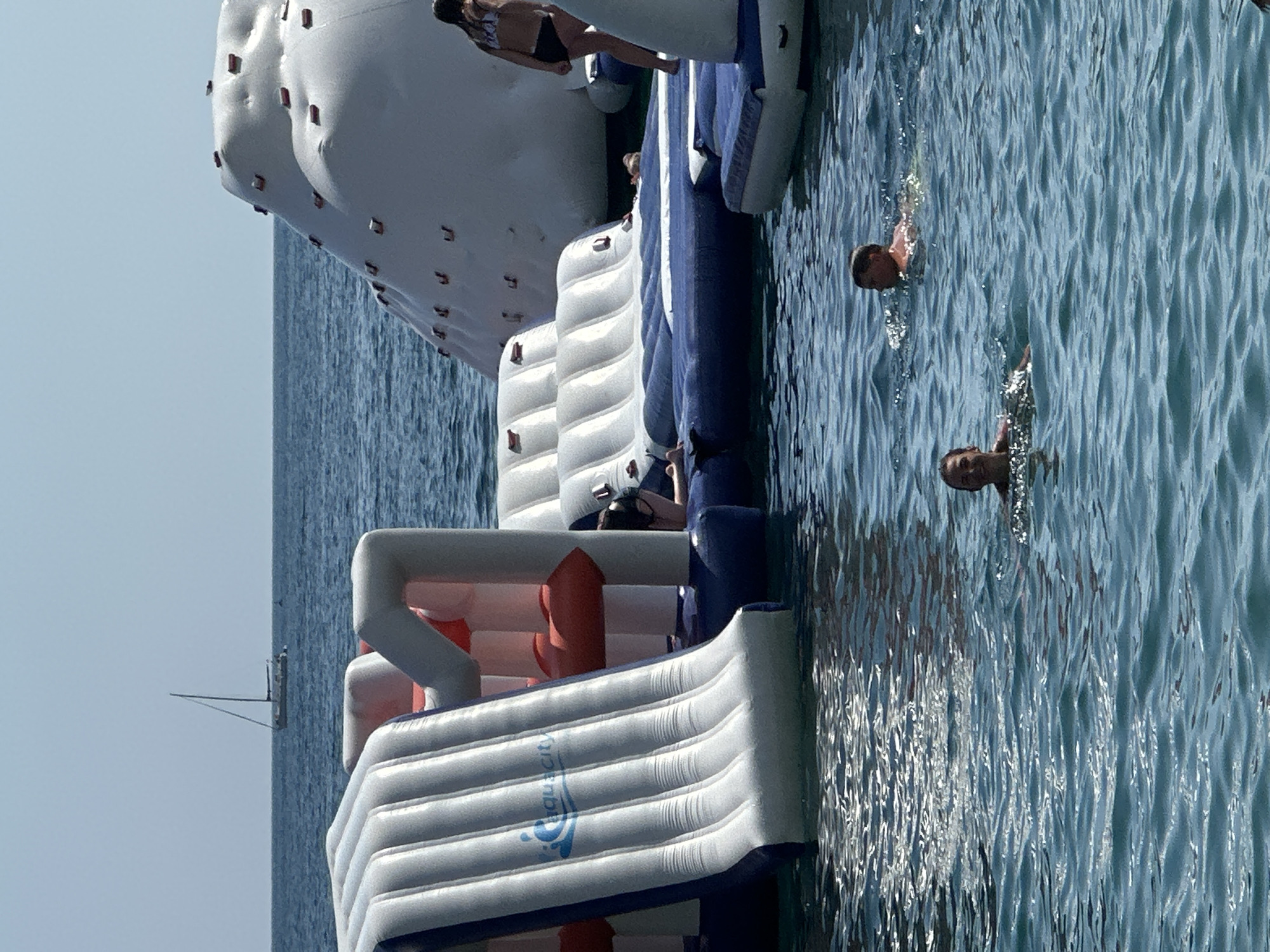 The image depicts a group of people enjoying an inflatable water park on a bright, sunny day. The water park is a large, multi-level structure made of white and blue inflatable modules, including slides, climbing walls, and platforms. In the foreground, the top of a large white inflatable slide is visible, with the "AquaCity" logo imprinted on it.
Several people are engaged in activities within the water park. One person is seen on the top platform of the inflatable structure, appearing to be preparing to jump. In the water, two individuals are swimming, their heads and shoulders visible above the surface. A person's hand is also visible reaching out from the water. The water itself is a clear blue, with gentle ripples reflecting the sunlight.
In the background, a vast expanse of calm, blue water stretches to the horizon under a clear, pale blue sky. A small sailboat with a single sail is visible in the distance, suggesting a coastal or open water setting. The overall atmosphere is one of leisure and recreation, with the bright sunlight and the presence of people enjoying themselves indicating a pleasant summer day. The image appears to be taken from a slightly elevated angle, looking down at the water park and the swimmers.