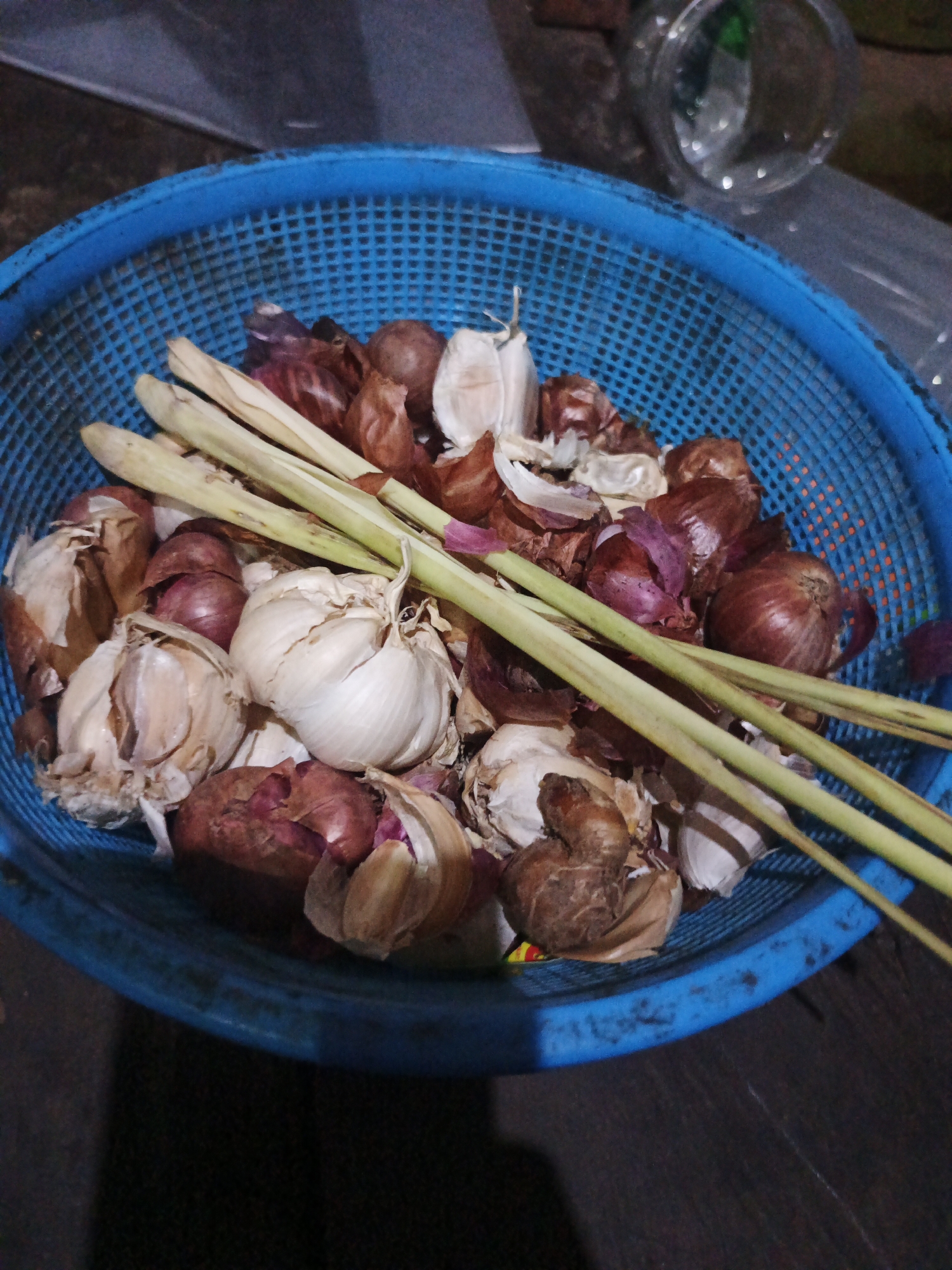 A blue plastic basket contains a mixture of ingredients: several bulbs of garlic, red onions, and stalks of lemongrass. The basket is placed on a dark, wooden surface. The lighting is dim, casting shadows. The background is blurred and indistinct, but appears to be an indoor setting, possibly a kitchen counter or table. There are no people or discernible actions. The scene is still and appears to be a collection of raw ingredients. The image is a close-up shot, focusing on the contents of the basket. There is no information within the image to suggest a specific location in Sinar Bulan, Indonesia.