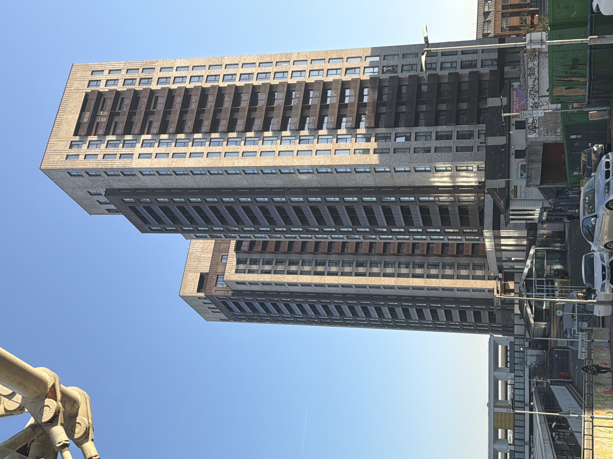 Here's a concise description of the image:
The image shows a tall, modern apartment building constructed of concrete, featuring regular rows of windows and projecting angled elements. It appears to be composed of at least two connected sections, differing slightly in height. A clear blue sky is above the building.
The base of a large metal structure is visible in the lower left. A street is on the right side of the image, with parked cars, people, and other buildings visible. Graffiti is present on some of the street-side structures.