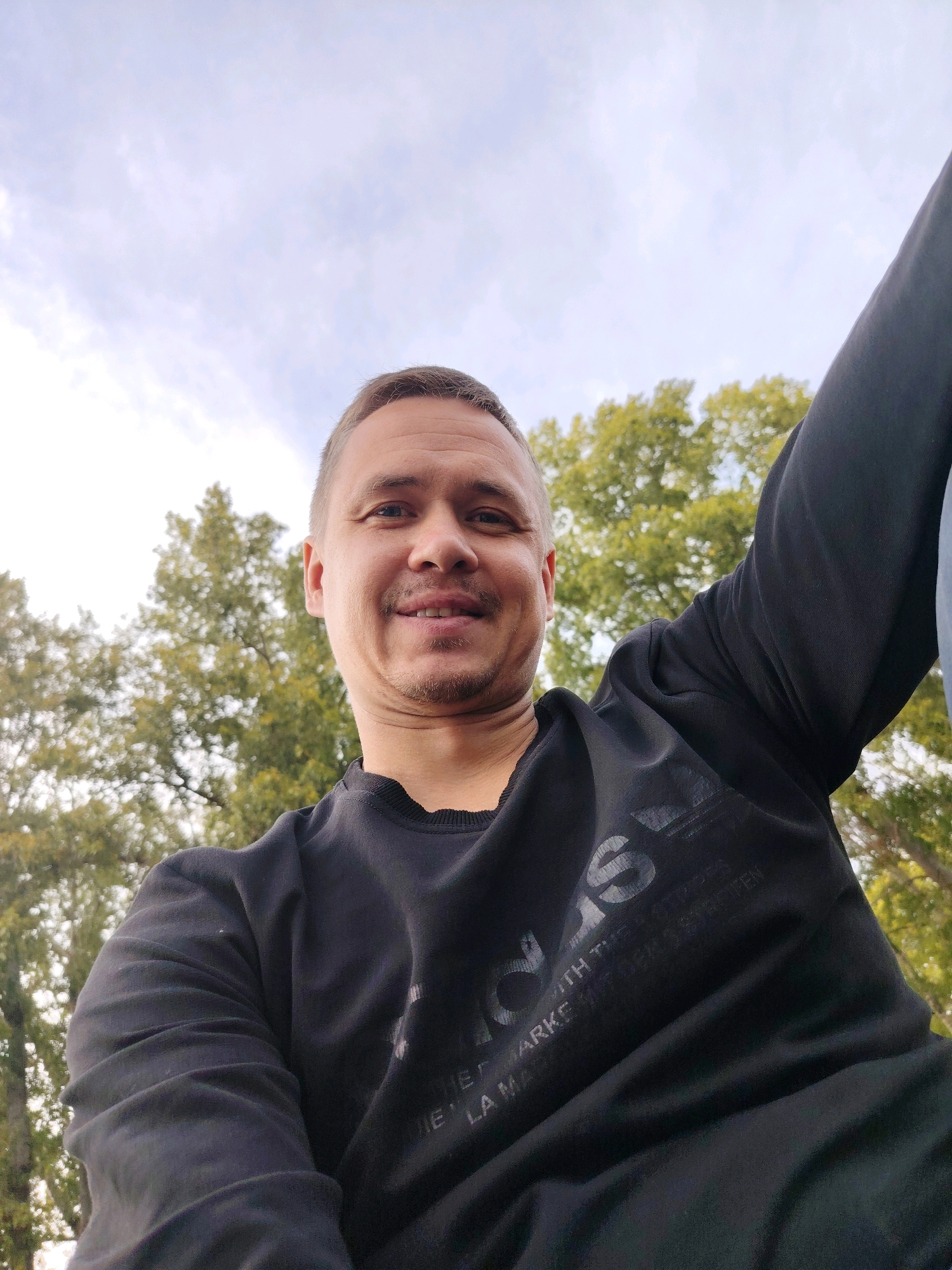 A low-angle, wide shot shows a light-skinned man with short, brown hair and a mustache smiling into the camera. He is wearing a black, long-sleeved shirt with a logo on the chest. The man is outdoors, with lush green trees and a cloudy sky visible in the background. The scene appears to be a park or natural area.
