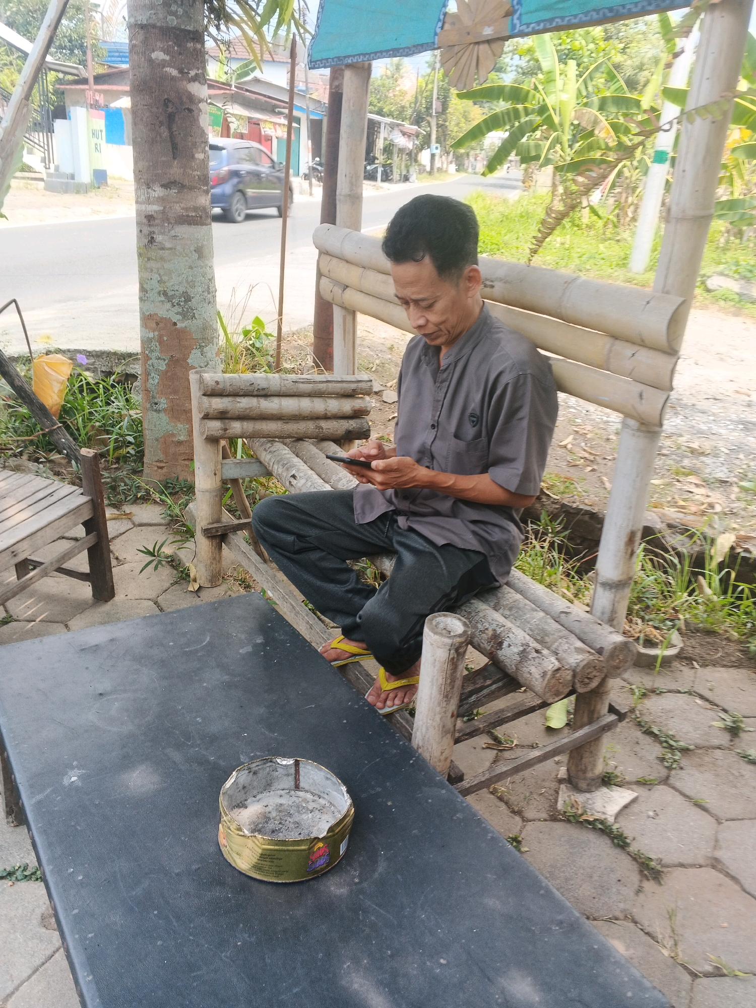 The image depicts an outdoor scene in Ungaran, Indonesia, likely near a roadside stall or rest area. In the foreground, a black table is visible, upon which rests a used ashtray containing cigarette butts and ash. To the left of the table, a wooden chair is partially visible.
The central subject is a middle-aged man of Asian descent, wearing a grey button-up shirt and dark trousers. He is seated on a rustic bamboo bench, his attention focused on a smartphone he holds in his hands. His posture suggests engagement with the device. The bamboo bench is weathered and constructed from thick bamboo poles.
Behind the man, a tree trunk with textured bark stands prominently. Beyond that, a road is visible with a blue car driving away from the camera. The background shows vegetation, including banana trees, suggesting a tropical or subtropical environment. Structures that appear to be small buildings or stalls are also present in the distance, hinting at a commercial or residential area alongside the road. The lighting indicates it is daytime, likely with bright sunlight. The overall ambiance is casual and appears to be a typical roadside setting in a town or city in Indonesia.