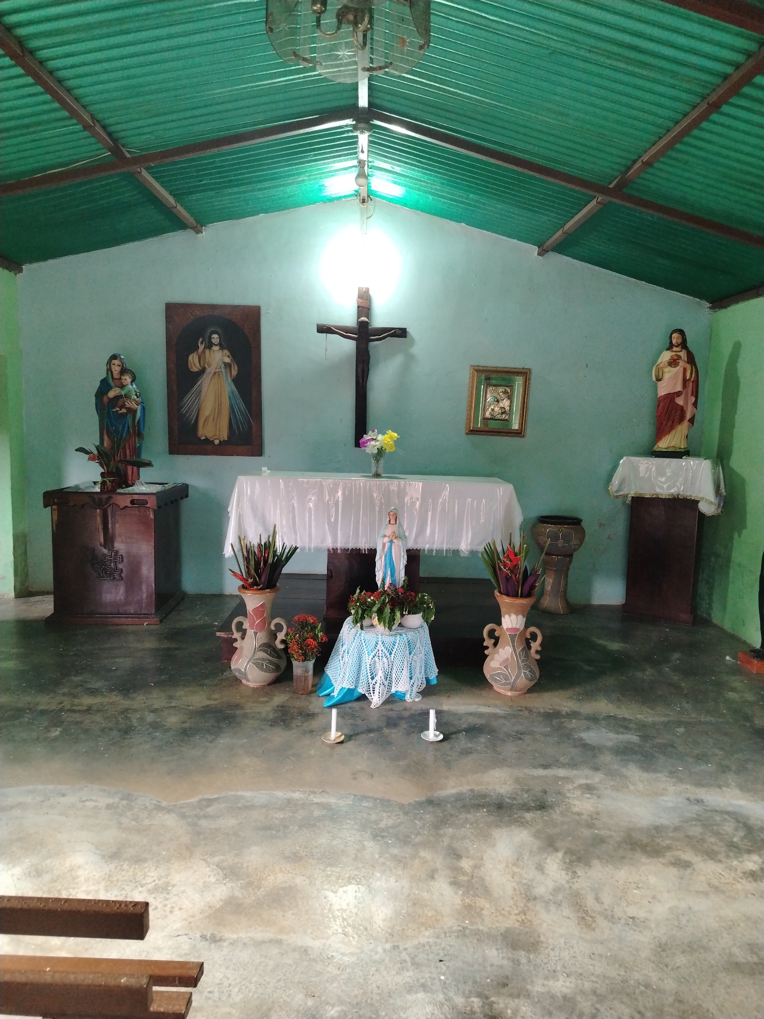 The image depicts a small, simple chapel in what appears to be Los Leoncitos, Venezuela. The building has a corrugated metal roof supported by exposed wooden beams. The walls are painted a light blue-green color.
Inside, the chapel contains several religious objects. On the left is a statue of Mary holding the infant Jesus atop a wooden pedestal. Next to it hangs a framed picture of Jesus. In the center, a table covered with a white cloth holds a crucifix and a vase of flowers. Below the table is a smaller table draped with a light blue crocheted cloth, topped with a statue of Mary surrounded by flowers. Two candles stand on the floor in front. Vases containing flowers flank this setup. On the right side of the chapel, there's a statue of Jesus atop a wooden pedestal. A framed picture also hangs on the wall. A drum sits between the alter table and the statue.
The floor is concrete and appears to be well-worn and possibly damp. A few wooden bench slats are visible in the foreground. The lighting is provided by an overhead light fixture, resulting in a bright spot on the back wall behind the crucifix. The overall impression is of a modest, well-used, and cared-for religious space.