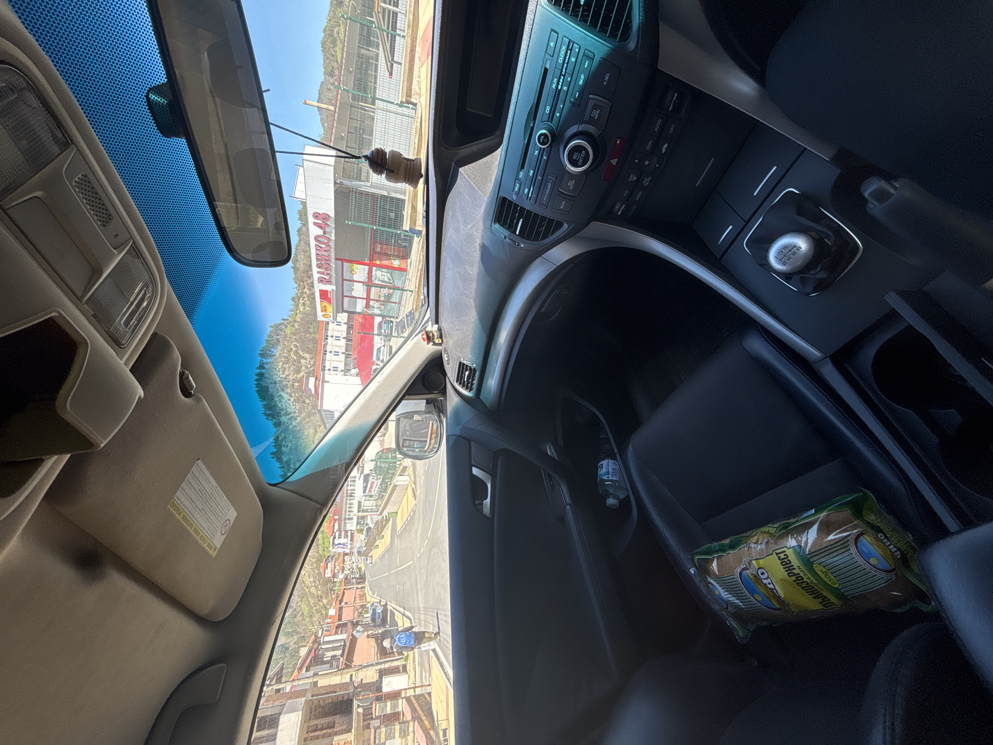 A car interior photograph taken in Zlatograd, Bulgaria, showcases a view through the windshield towards Rashko-48, a local business with red and white signage. The interior includes a rearview mirror, a dashboard with a car radio unit, a manual transmission shift knob, and a visible bag of 'Глълнозърнест' crackers. A plastic bottle is in the door, and the road outside is lined with buildings. The sun visor is down.
