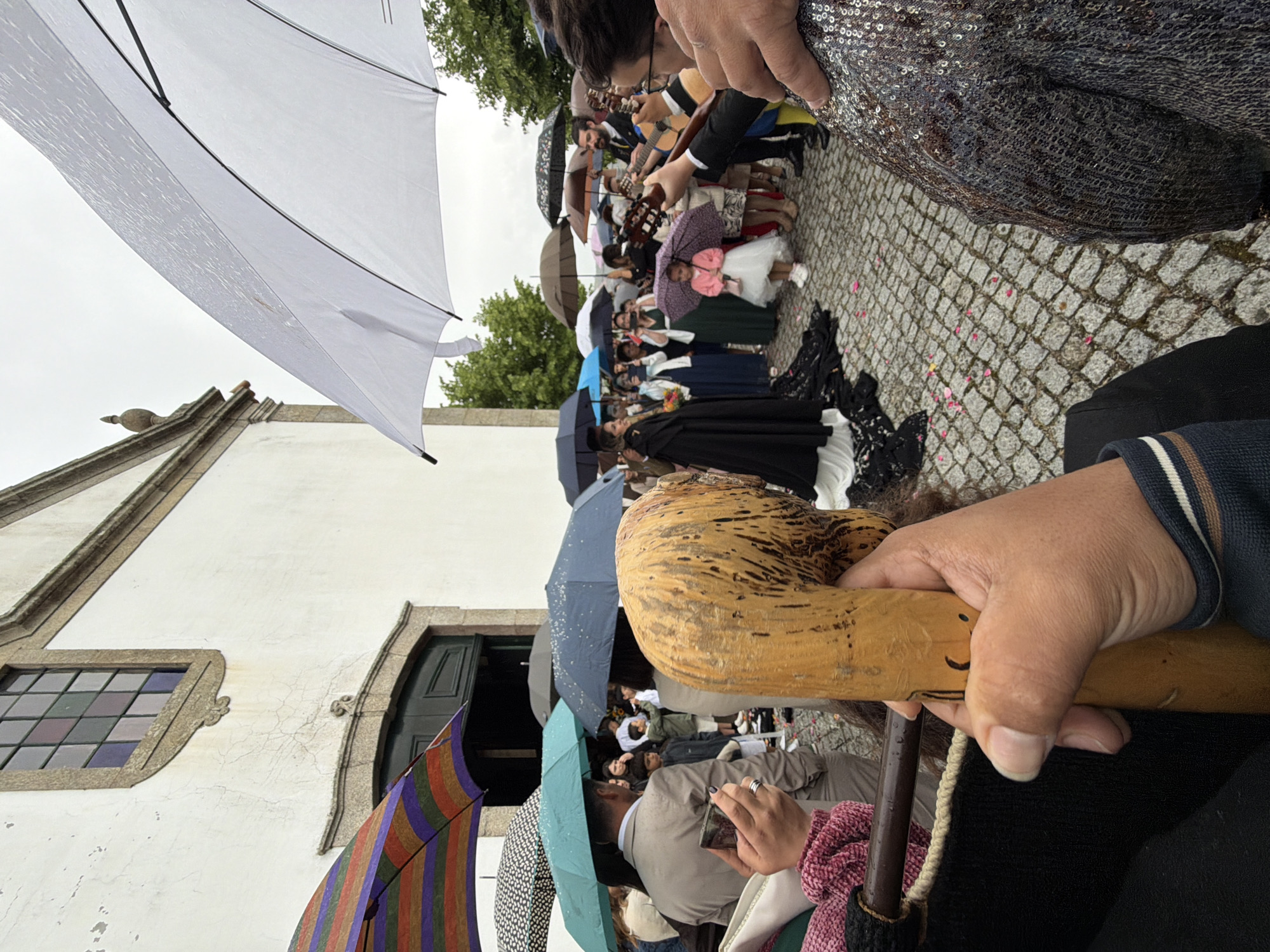 A crowd of people holding umbrellas are standing outside a white building with a stained-glass window and open green doors, possibly a church. Some individuals are playing instruments, including guitars. There are flower petals scattered on the cobblestone pavement. The presence of umbrellas suggests it is raining or has recently rained. The location is Vila Real, Portugal.