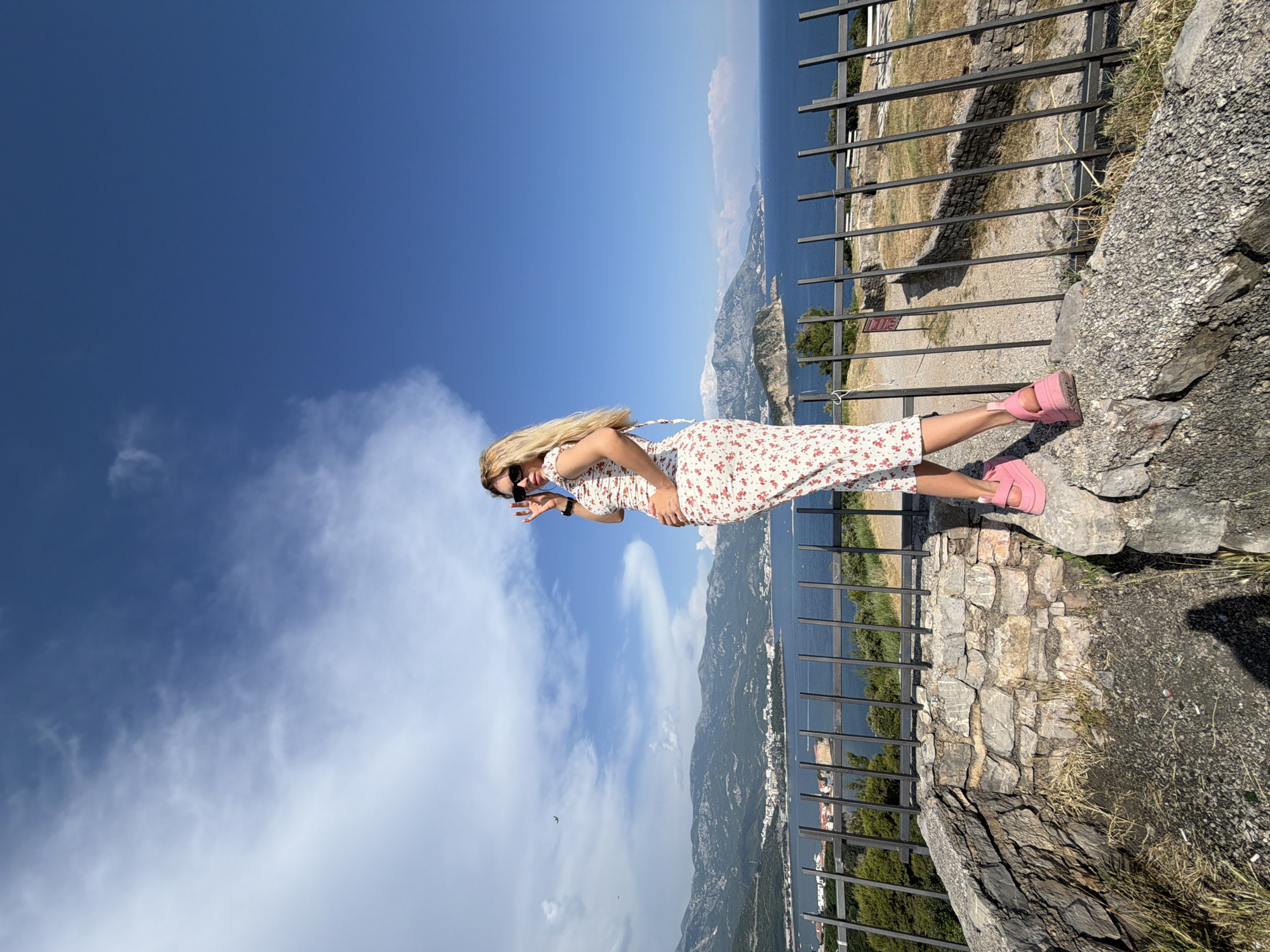 A woman stands on a stone embankment overlooking the Adriatic Sea and the city of Budva, Montenegro. She has blonde hair and wears a floral print dress and pink platform shoes. She is looking over her shoulder with her hand on her hip and sunglasses on her face. The sky is blue with scattered clouds, and mountains rise in the background. A black metal fence separates the embankment from the view.