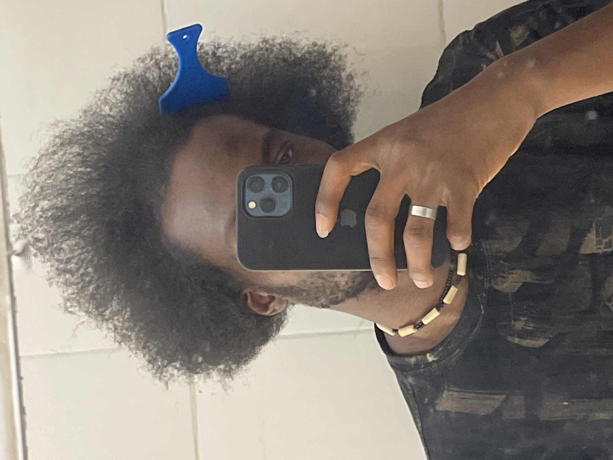 A person is holding a dark-colored smartphone in front of their face, likely taking a selfie in a bathroom. They have dark curly hair with a blue comb in it. The person is wearing a dark patterned shirt and a beaded necklace. A silver ring is on one of their fingers. The background shows a white tiled wall, indicative of a bathroom. The location is unknown.