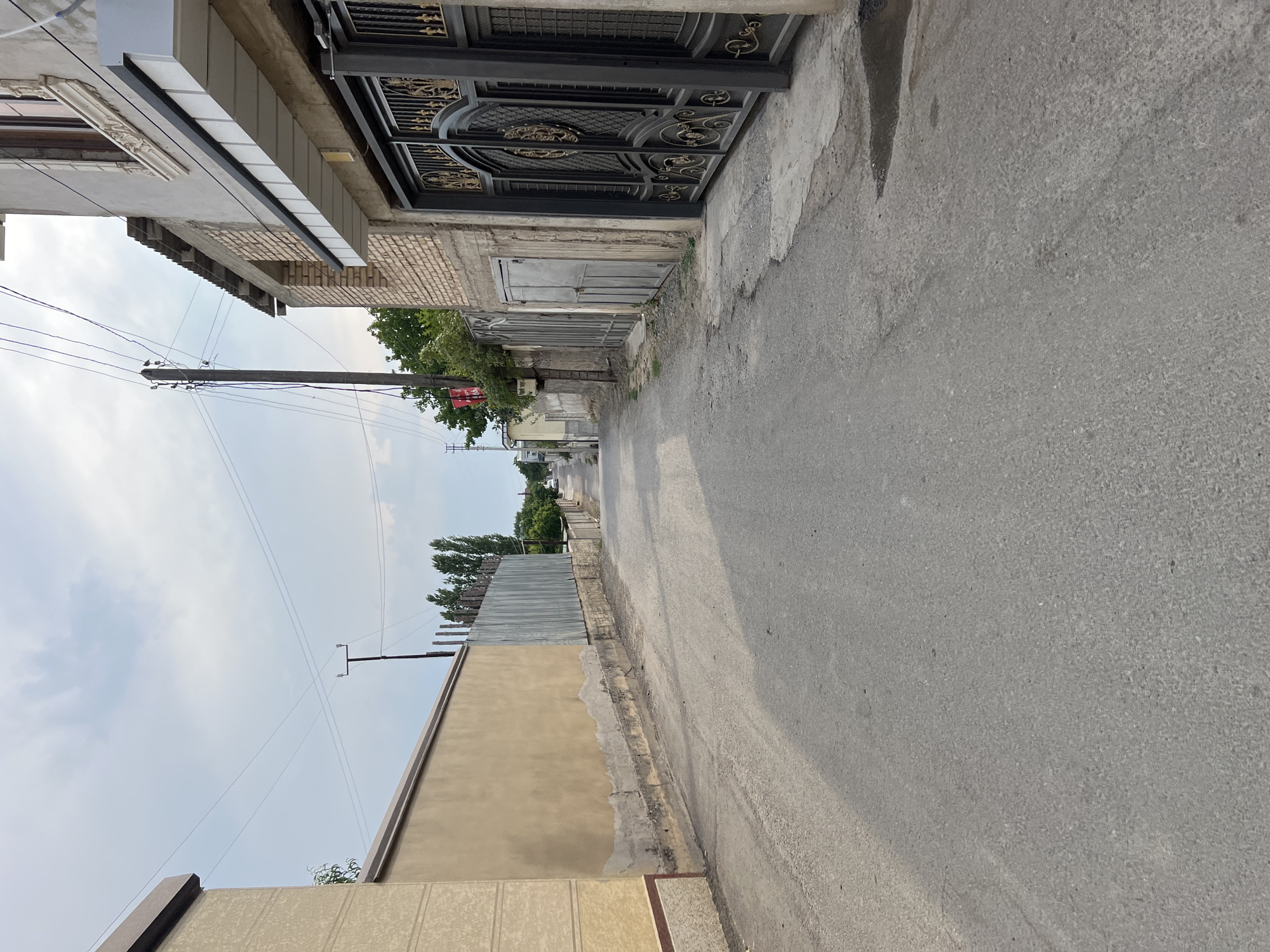 The image shows a narrow, asphalt road in a residential area, likely in the city of Guliston, Tajikistan. The road is lined with walls and buildings on both sides. On the left, there is a tan-colored wall with a textured finish, followed by a corrugated metal fence. On the right, a building with a light-colored facade is visible, featuring a large, ornate black gate with gold detailing. Further down the street, more buildings and fences can be seen, along with trees and utility poles with power lines. The sky is partly cloudy. The overall impression is of a quiet, somewhat worn urban streetscape.