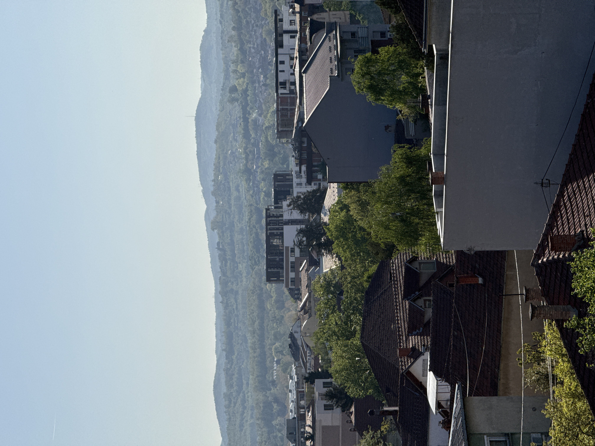 This image presents a view of Kragujevac, Serbia, from an elevated position. In the background, there is a hazy mountain range. Mid-ground features residential buildings and interspersed greenery, typical of a suburban setting. Foreground elements consist of building rooftops with terracotta tiles and chimneys. There are no visible people or distinct actions. The environmental conditions indicate a clear, possibly sunny day due to the brightness and lack of precipitation.