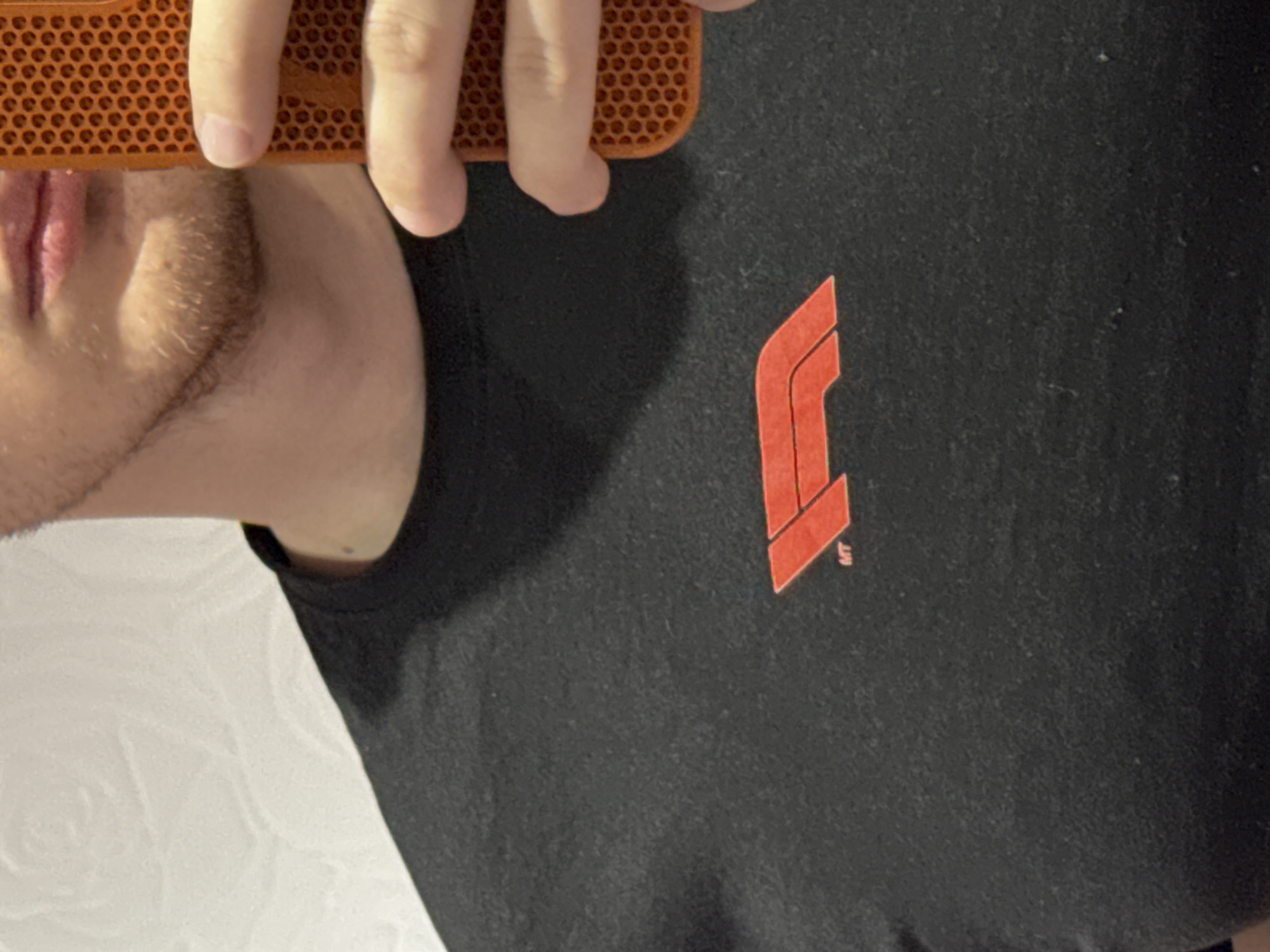 The image shows a person wearing a black t-shirt with the Formula 1 logo in red. A portion of the person's face, specifically the mouth and facial hair, is visible, partially obscured by an orange object held up to the camera. The background appears to be a white, textured wall. There is no information indicating the location is Plovdiv, Bulgaria.