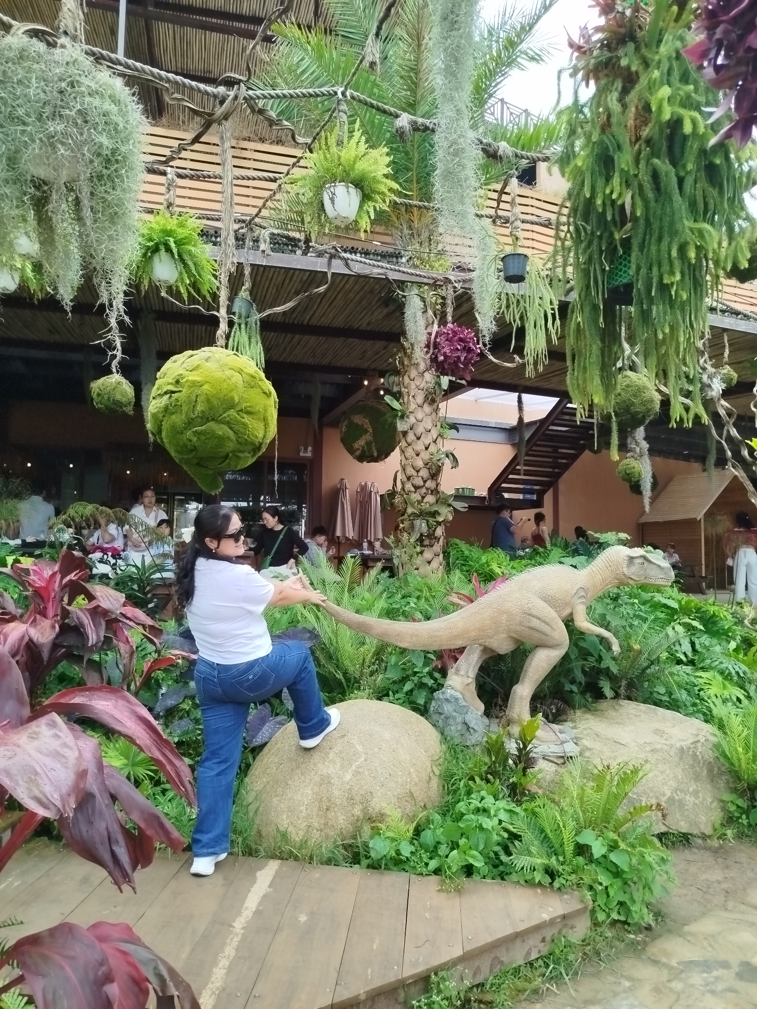 A woman is posing in front of a dinosaur statue in a garden in Lạc Dương, Vietnam. She is wearing a white t-shirt, blue jeans, and white sneakers, and is leaning on a rock with her left leg raised. The dinosaur statue is a light brown, standing on a rock surrounded by lush green plants. In the background, there are people dining at tables and a building with a wooden roof. Hanging plants and large, moss-covered spheres are suspended from the ceiling of the covered outdoor area. The scene appears to be a restaurant or cafe with a unique, naturalistic theme.