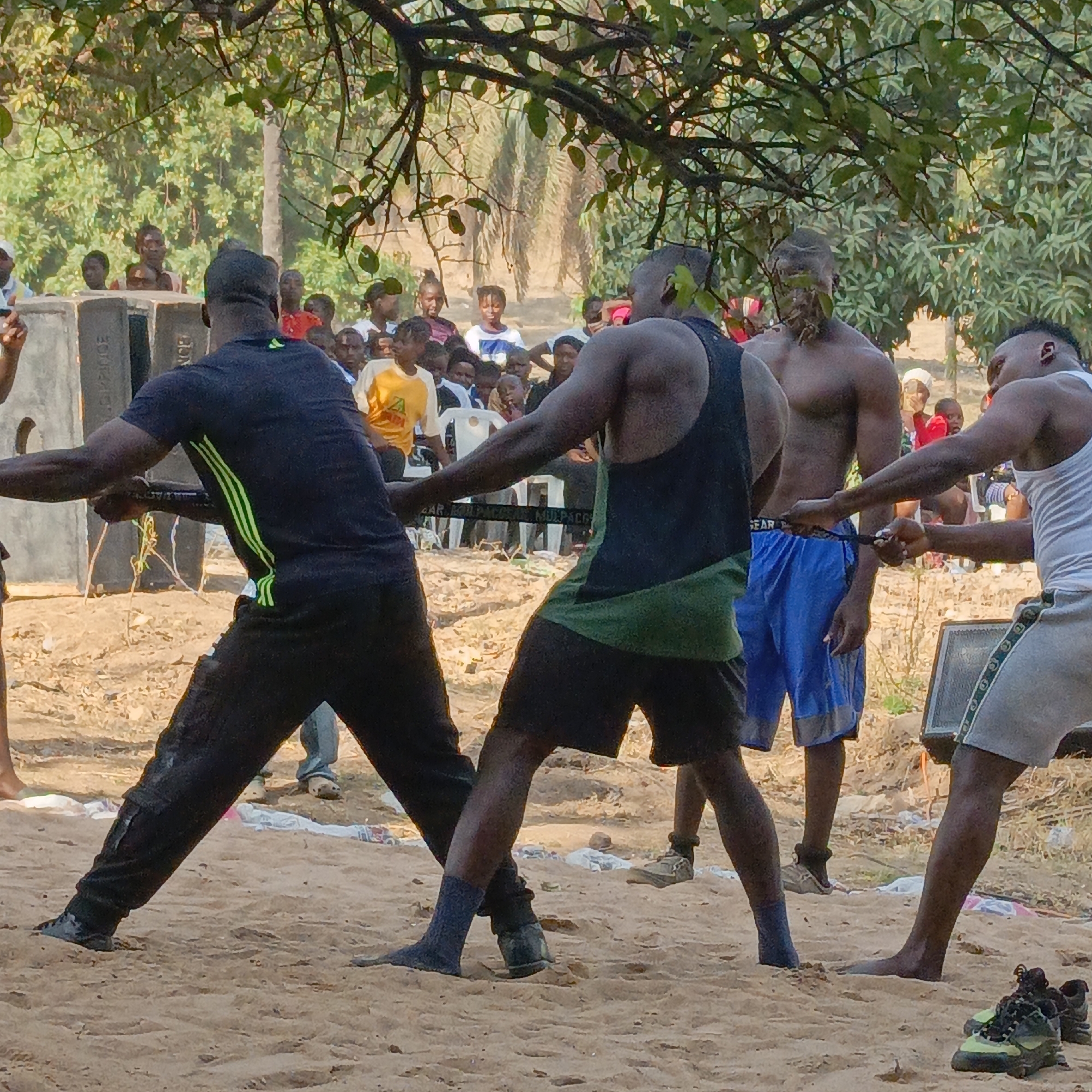 The scene depicts an outdoor event in Akwanga, Nigeria, featuring a physical competition resembling a tug-of-war. Three individuals are actively pulling a black strap across a light brown, sandy dirt ground.
On the left, a black male wears a black short-sleeved shirt with vertical green stripes on the back and black pants; his visible left foot is clad in a dark sock and dug into the sand. Adjacent to him, a second black male, with a muscular build, wears a black and dark green tank top and dark shorts; his feet are in dark socks. Both individuals are leaning forward, pulling the strap with both hands.
To the right, a third black male, also muscular, wears a white striped tank top and light grey shorts with a dark green side stripe; his feet are bare. He is leaning back, pulling the strap with both hands. A small lesion is visible on the back of his head near the hairline. A pair of athletic shoes rests on the ground near his bare feet.
Behind the central participants, a fourth black male, bare-chested and wearing blue shorts, stands observing the activity.
The black strap being pulled features partial white text, readable as "SAR KILOPACK MU.PACG".
In the background, a diverse crowd of spectators, consisting of men, women, and children of various ages, observes the event. Some individuals in the crowd are seated on white plastic chairs. Two large, rectangular, grey speaker boxes are positioned on the left side of the scene, one bearing the vertical text "DANCE" and "SOUND". Another similar speaker box is partially visible on the right.
The environment is characterized by abundant green foliage and tree branches overhead, casting shadows and suggesting a sunny day. The ground is uneven, with scattered white and colored debris. Dense vegetation forms the distant background.