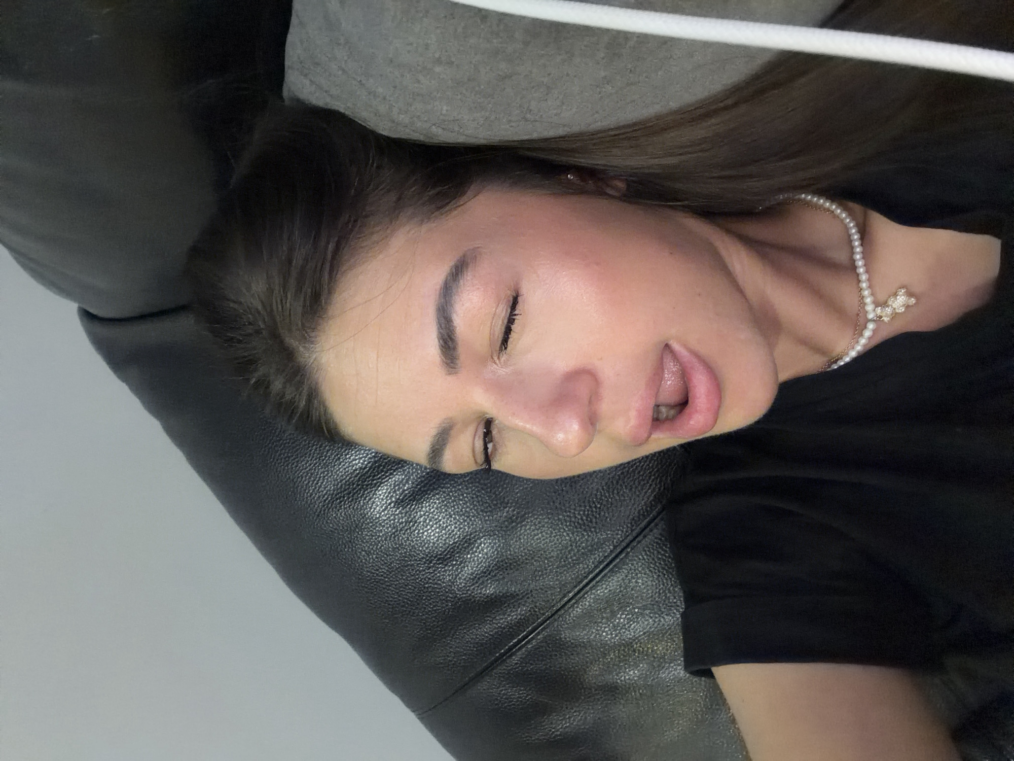 A young woman with brown hair is lying on a black leather couch, with a grey cushion behind her head. She is wearing a black t-shirt and a pearl necklace with a teddy bear pendant. Her right eye is closed, and her mouth is open, with her tongue slightly out. The background is a plain grey wall. The image appears to be taken indoors. No specific location in Novi Iskar, Bulgaria is identifiable.