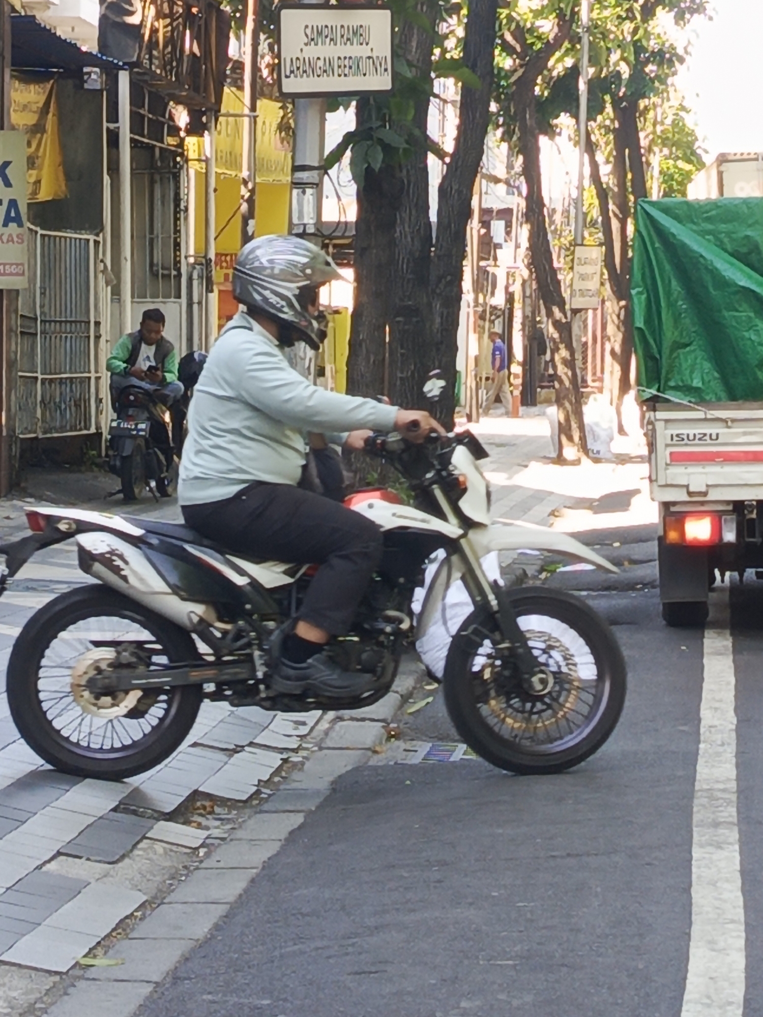In Surabaya, Indonesia, a man wearing a light blue long-sleeved shirt, black pants, and a helmet is riding a white and black dirt bike. He is on a street with a sidewalk made of square pavers. The street is paved with asphalt and has a white lane marker. To the right of the motorcycle, a white truck with the "ISUZU" logo is visible. The truck has a green tarp covering its cargo. In the background, trees line the street, and there are buildings on the left side. A sign that reads "SAMPAI RAMBU LARANGAN BERIKUTNYA" is visible on a pole. Another man wearing a green jacket is sitting on a motorcycle in the background. The lighting suggests it is daytime, possibly late morning or early afternoon, with sunlight casting shadows.