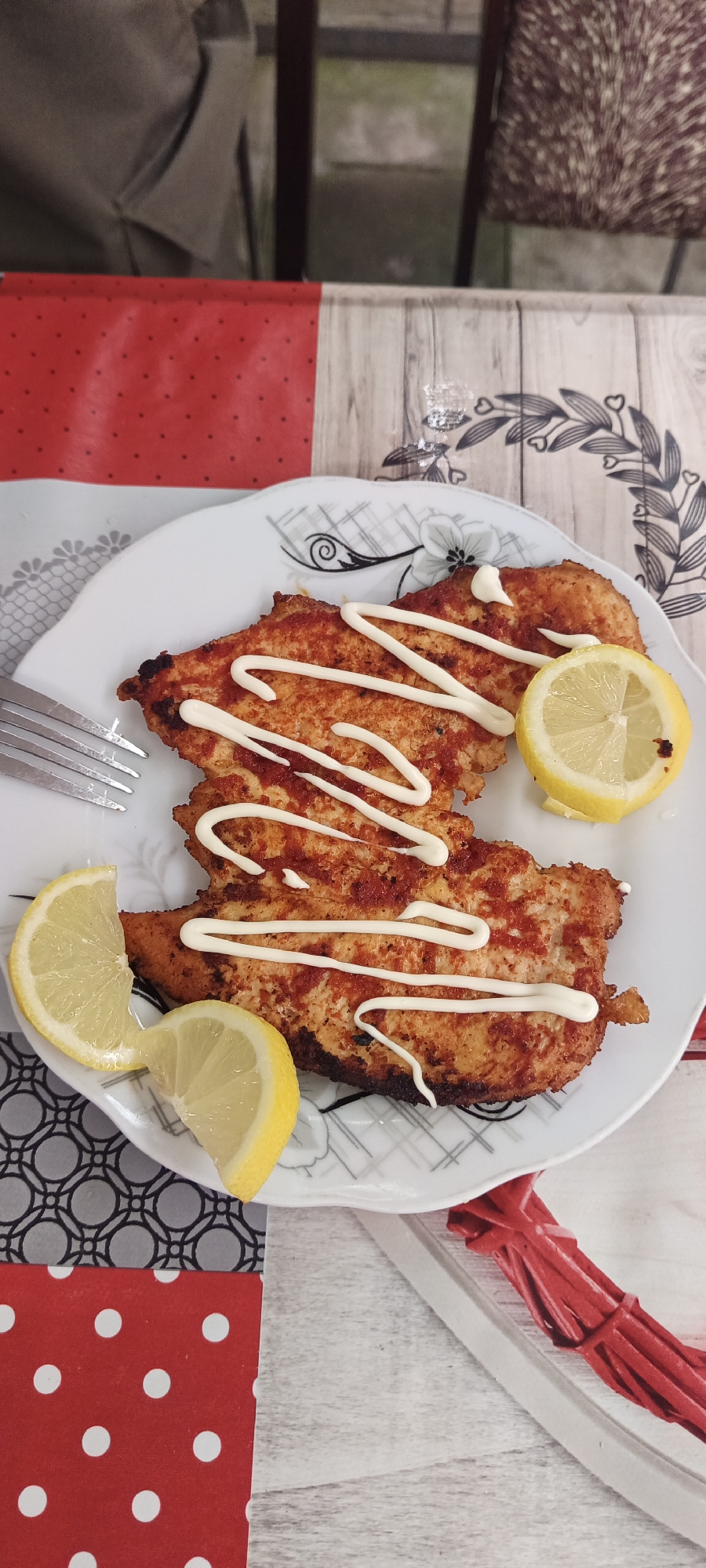 A plate of three breaded chicken cutlets is presented on a white plate with a decorative black and white floral pattern. The cutlets are drizzled with a white sauce, possibly mayonnaise, in a zigzag pattern. Two lemon slices are placed on the plate, one to the left and one to the right of the chicken. A fork is visible to the left of the plate. The plate sits on a table covered with a red placemat with white polka dots on the left, and a light wood-grain patterned surface on the right. The background shows hints of furniture and possibly a window. There is no indication of the location within the city of Khudat, Azerbaijan.