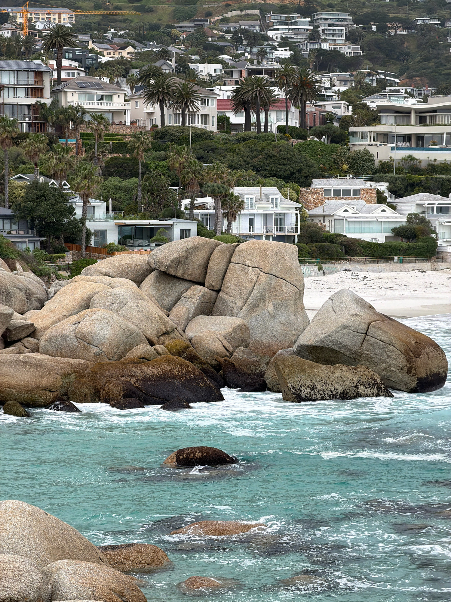 This image depicts a coastal scene in Camps Bay, Cape Town, South Africa. In the foreground, large, weathered granite boulders are partially submerged in the turquoise ocean, with white waves crashing around them. Beyond the boulders, a sandy beach is visible, leading up to a hillside densely populated with luxurious white houses, many featuring large windows, balconies, and flat roofs. These homes are nestled amongst lush green vegetation and palm trees, creating a picturesque residential area on the slopes. In the background, the hills of the city rise, also dotted with more residences. The overall impression is one of affluent coastal living in a vibrant, hilly city.