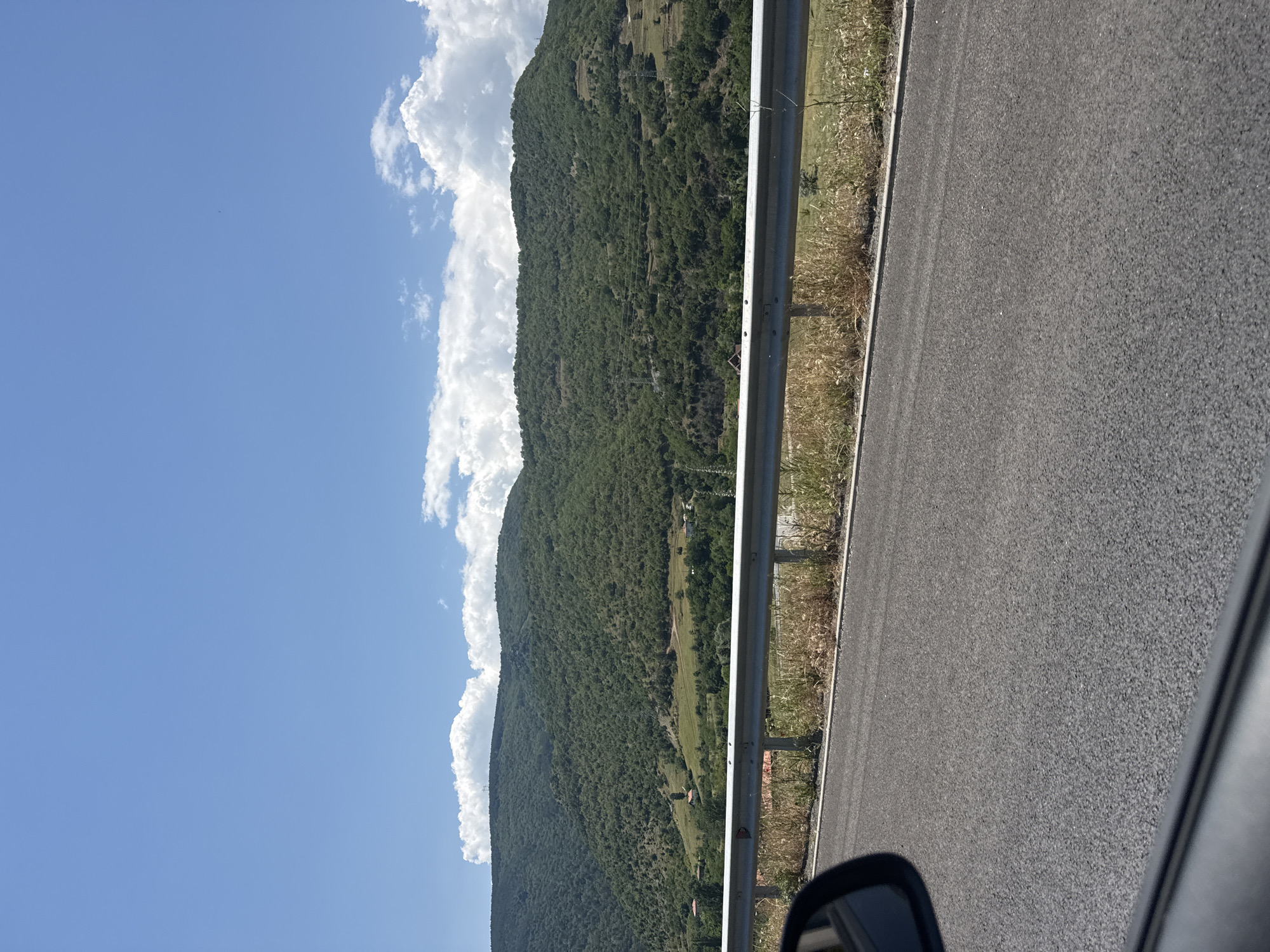 The image presents a panoramic view of a mountainous landscape from the perspective of a vehicle, likely taken in Jeleznitsa, Bulgaria. The scene is dominated by a clear blue sky and lush green mountains.
In the foreground, a dark asphalt road occupies the right side of the frame, indicating the direction of travel. A silver metal guardrail runs parallel to the road, separating it from a strip of dry, light-colored grass and weeds. A portion of a car's side mirror is visible in the bottom right corner, reinforcing the viewpoint from a moving vehicle.
The midground and background feature a majestic mountain range, heavily covered with dense green trees and foliage. Patches of terraced land or clearings are discernible on the mountain slopes, along with a few scattered small buildings, possibly houses or farm structures. Faint lines, possibly power lines, can be seen traversing parts of the mountainside.
The sky above the mountains is a vibrant blue, filled with large, fluffy white cumulus clouds that appear to rest atop the mountain peaks. The bright natural light suggests it is daytime, likely a sunny morning or afternoon. There are no people visible in the image, nor any discernible text. The overall atmosphere is one of serene natural beauty and travel through a picturesque landscape.