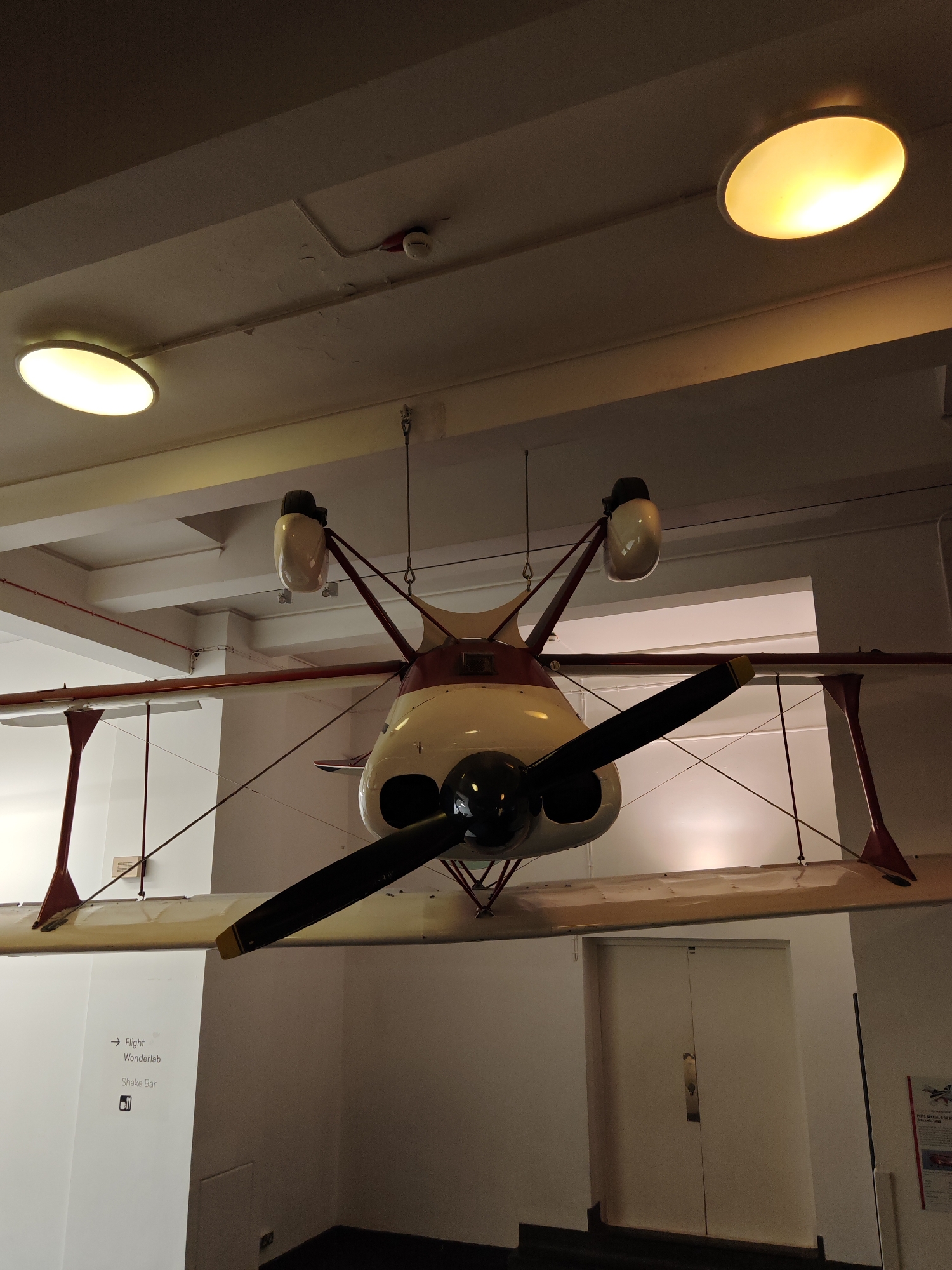 A vintage aircraft, likely a biplane, is suspended from the ceiling of a building, possibly a museum or exhibit space, in London, United Kingdom. The aircraft is painted in a combination of white, red, and black, with a prominent propeller at the front. The ceiling is white and features two round, illuminated light fixtures. A sign indicating directions to "Flight Wonderlab" and "Shake Bar" is visible on a nearby wall. A closed, white double door is in the background. No people are visible in the frame.