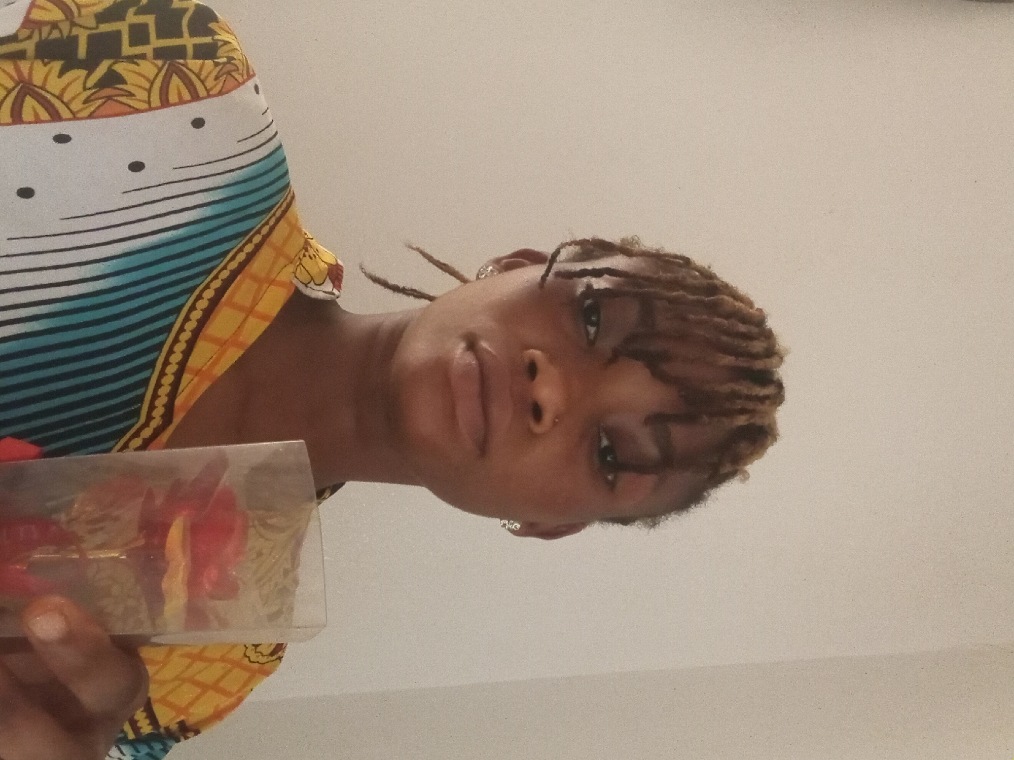 A woman with light brown dreadlocks is holding a clear plastic box in her left hand. The box contains red and yellow objects. She is wearing a colorful patterned garment with white, yellow, and blue stripes. The background is a plain, light-colored wall. The photo appears to be taken from a low angle. The location is unknown.