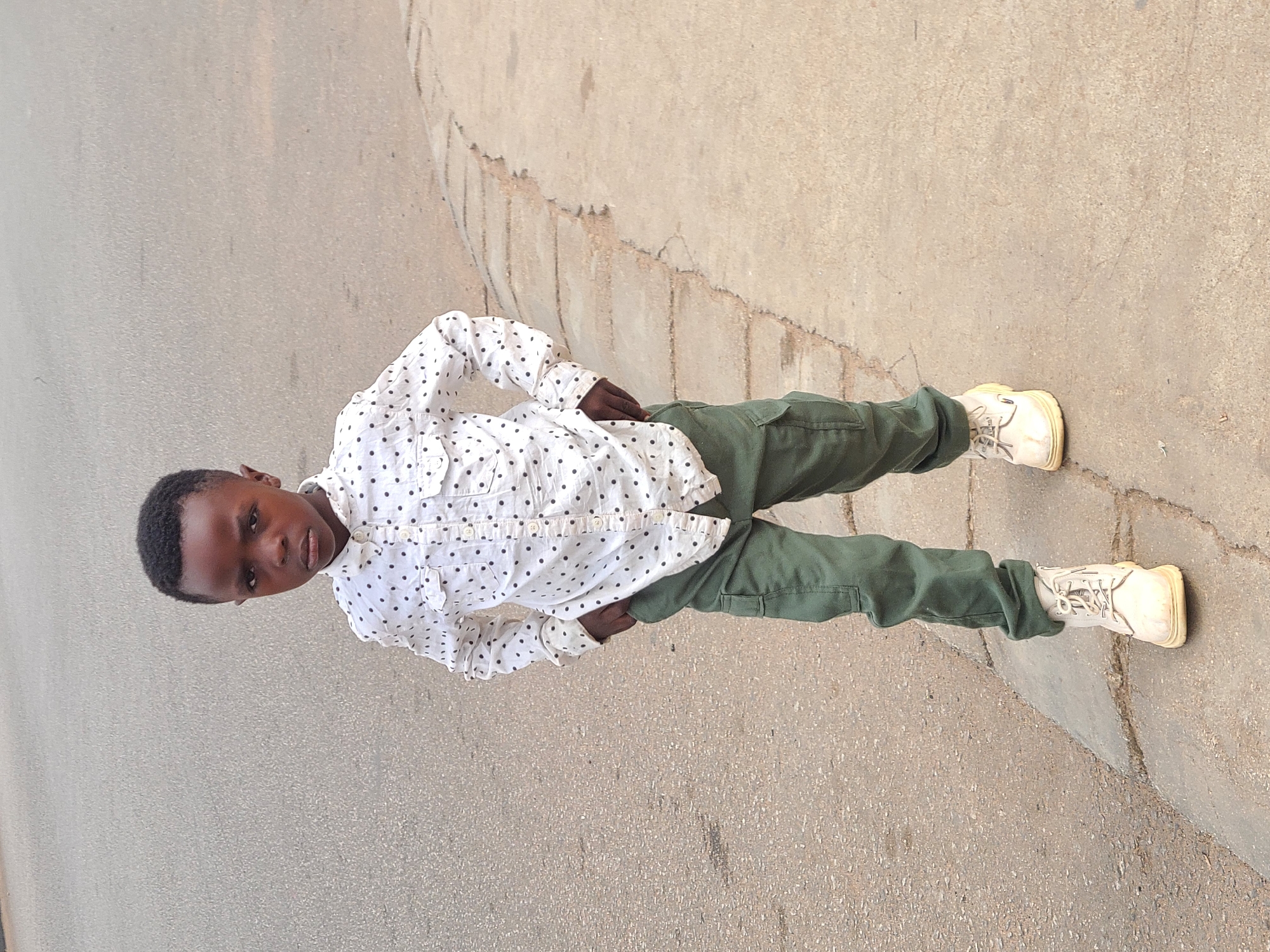 A young boy stands on a paved surface, likely a sidewalk or street in Jos, Nigeria. He is wearing a white, long-sleeved shirt with black polka dots, green pants, and off-white sneakers. He has his hands on his hips. The pavement surrounding him is cracked and shows signs of wear. The lighting suggests daytime.