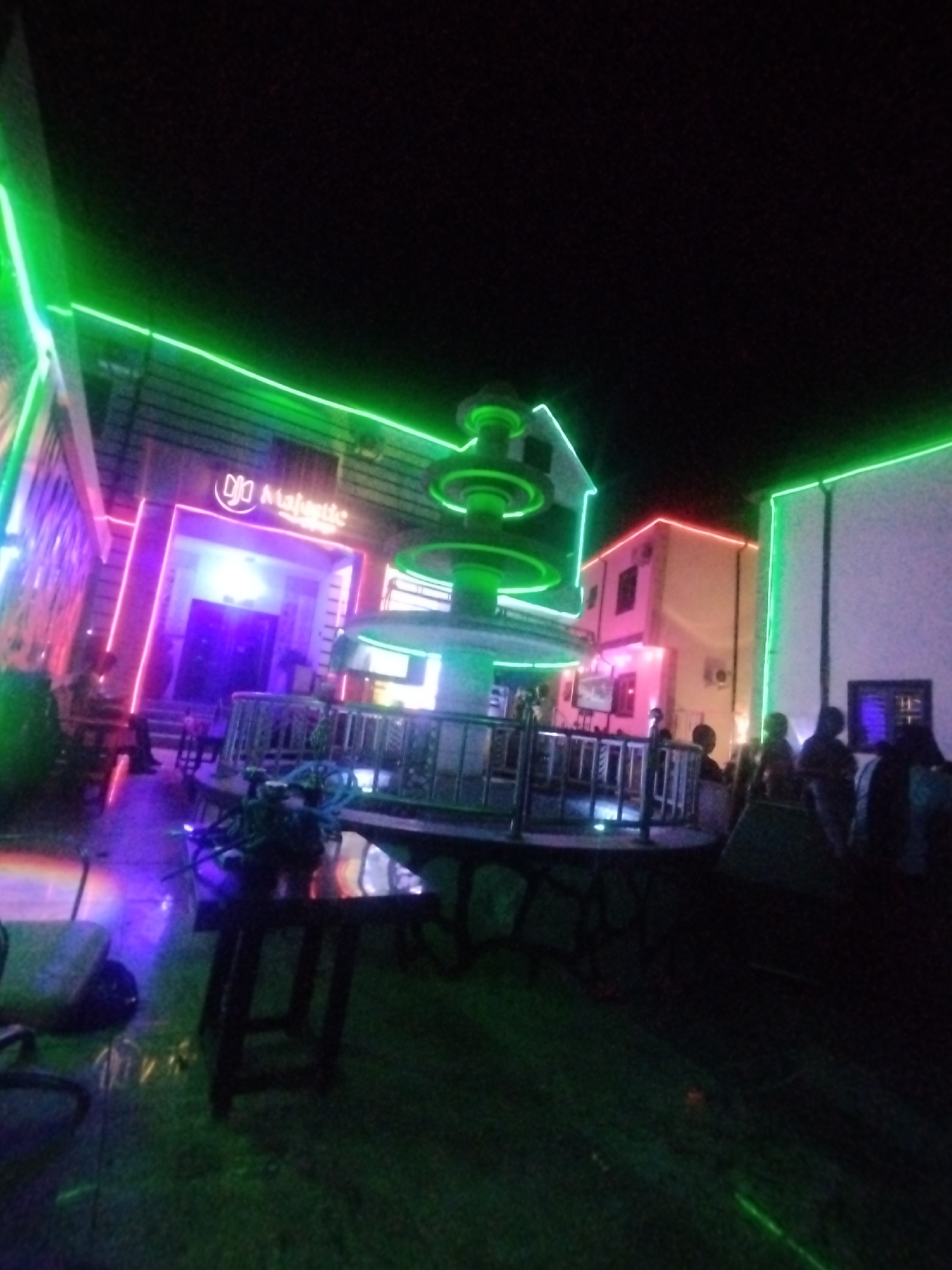The image depicts an outdoor area at night in Tumfure, Nigeria, featuring illuminated structures. The "Majestic" building is visible, outlined with purple and green neon lights, with a fountain illuminated with green neon lights in front. Additional buildings are visible, also outlined with neon lights. Several people are gathered in the background. Foreground includes tables and chairs. The ground appears to be a combination of paved and grassy surfaces.