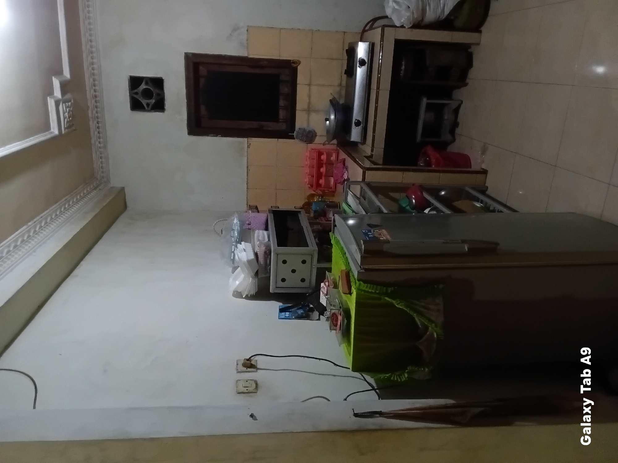The image depicts a simple, somewhat cluttered kitchen interior, likely within a residential setting in Trenggalek, Indonesia. The walls are painted a pale, off-white color, and the ceiling features decorative molding. A small, dark-framed window with an intricate geometric vent is visible on the right wall.
In the foreground, a large, gray refrigerator is partially visible, covered with a green fabric tablecloth that drapes over its top and sides. Various items, including what appear to be food containers and condiments, are placed on top of the tablecloth. To the right of the refrigerator, a small countertop area is set up with a two-burner gas stove, upon which a cooking pot rests. A red plastic container, possibly a trash can, is situated beneath the countertop. Shelving units are present, holding containers, bottles, and other kitchenware. A sink area is also visible with various cleaning supplies.
Electrical outlets and cords are present on the wall to the left, indicating the active use of appliances. The floor is tiled with light-colored square tiles. The overall impression is of a functional but modest kitchen space. The lighting appears to be artificial, creating some shadows and contributing to the slightly dim ambiance of the room. The text "Galaxy Tab A9" is superimposed in the bottom left corner of the image.