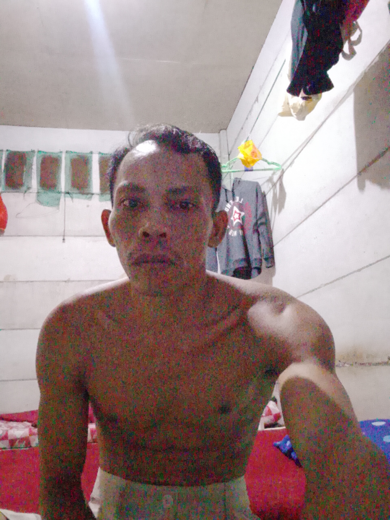 A dimly lit, indoor scene in Bukit Ketok, Indonesia, shows a shirtless man with short dark hair, light brown skin, and a neutral expression. He is partially visible from the chest up, facing the camera, and holding his arm out in a possible selfie pose. He is wearing light-colored bottoms. The background consists of white walls with a clothesline holding various items, including clothing and a yellow package on a hanger. The lower portion of the image contains bedding with different patterns. The overall image quality is grainy.