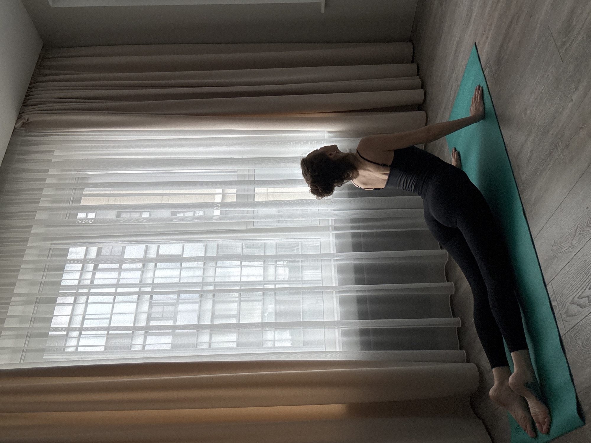 In a room in Khimki, Russia, a woman is practicing yoga on a teal mat. She is wearing a black bodysuit and her legs are extended behind her, with her feet resting on the mat. Her arms are bent, with her palms flat on the mat beside her chest, supporting her upper body as she arches her back upwards. Her head is turned to her left, looking towards a large window covered by sheer white curtains and heavier beige curtains. Through the sheer curtains, a building with a grid-like pattern of windows is visible. The floor is made of light-colored wooden planks.
