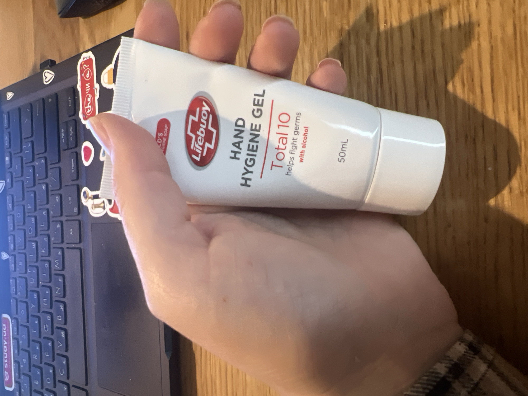 In Kyiv, Ukraine, a hand holds a small white tube of Lifebuoy Hand Hygiene Gel, labeled "Total 10 helps fight germs with alcohol," with a capacity of 50ml. The tube is positioned over a portion of a laptop keyboard, which is partially visible. The background consists of a wooden surface. Stickers are affixed to the laptop, including one with the text "study.ua". The scene is illuminated, casting a shadow of the hand and tube onto the wooden surface.