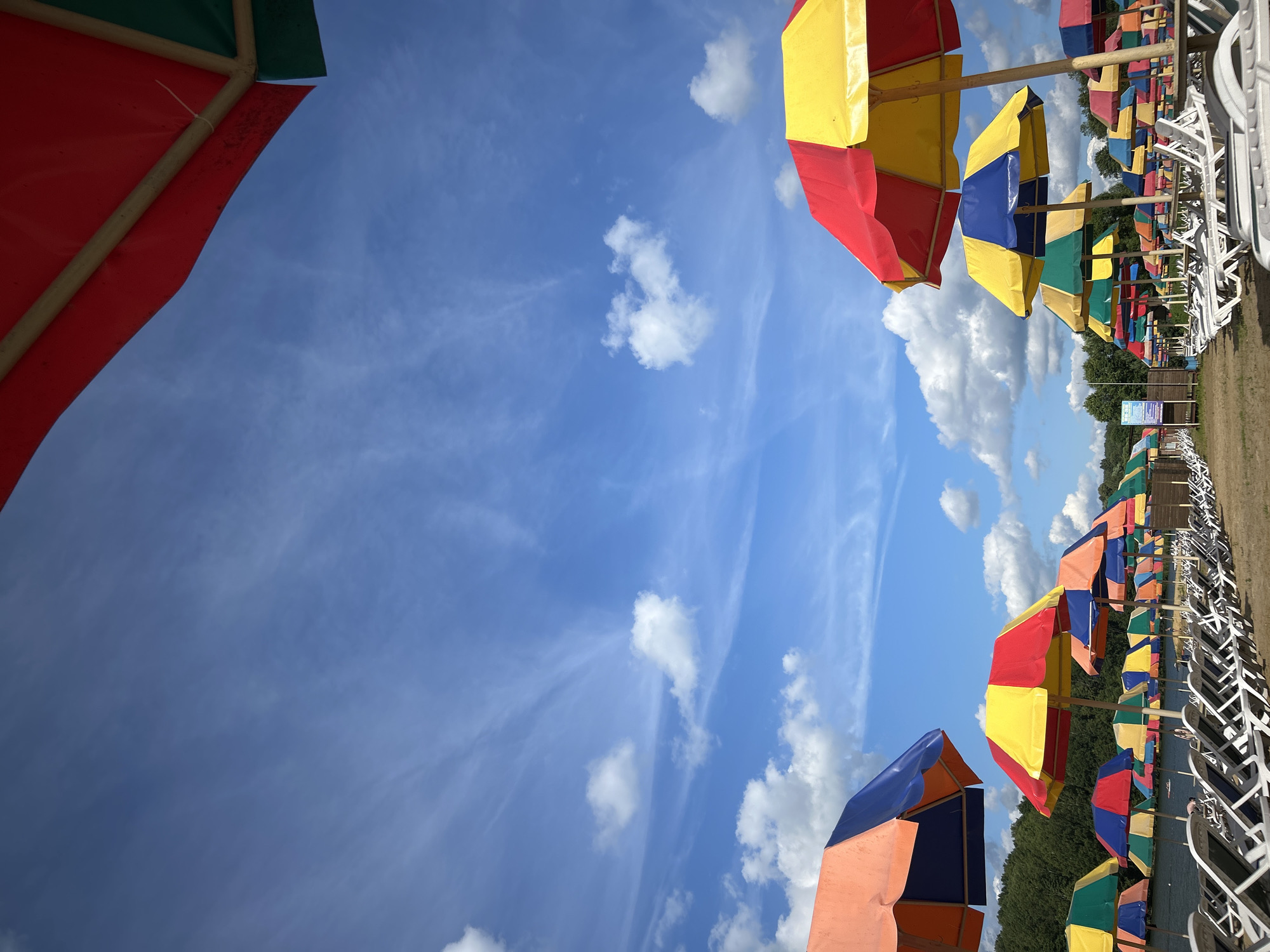 The image depicts an outdoor scene at a beach or recreational area, likely in the city of Yubileiny, Russia, given the prompt's reference. The foreground is dominated by rows of white lounge chairs arranged on sandy ground. Behind the chairs, a multitude of colorful beach umbrellas are set up. These umbrellas are multi-colored, featuring combinations of red, yellow, blue, and green panels.
In the background, a body of water, possibly a lake or river, is visible, bordered by a line of trees. The sky above is a clear blue, punctuated by scattered white clouds, some of which are wispy and others more substantial. The overall impression is one of a bright, sunny day at a leisure facility, prepared for visitors. No people are clearly visible in the image.