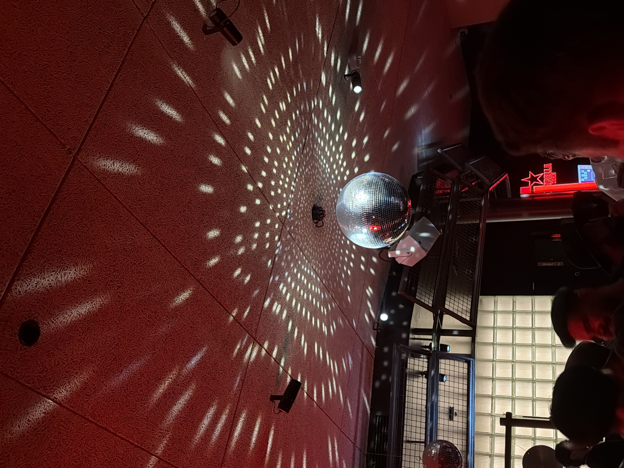 A tilted medium shot captures an indoor scene dominated by a mirrored disco ball suspended from the ceiling, casting light reflections across a red wall. Various track lights are affixed to the wall. To the right of the disco ball, a metal structure stands against a wall composed of glass blocks. The head of an individual is partially visible in the upper right corner. The setting is possibly a bar or nightclub located in Barcelona, Spain, judging by the red Estrella beer sign. The scene appears dimly lit, with the disco ball acting as the primary source of illumination.
