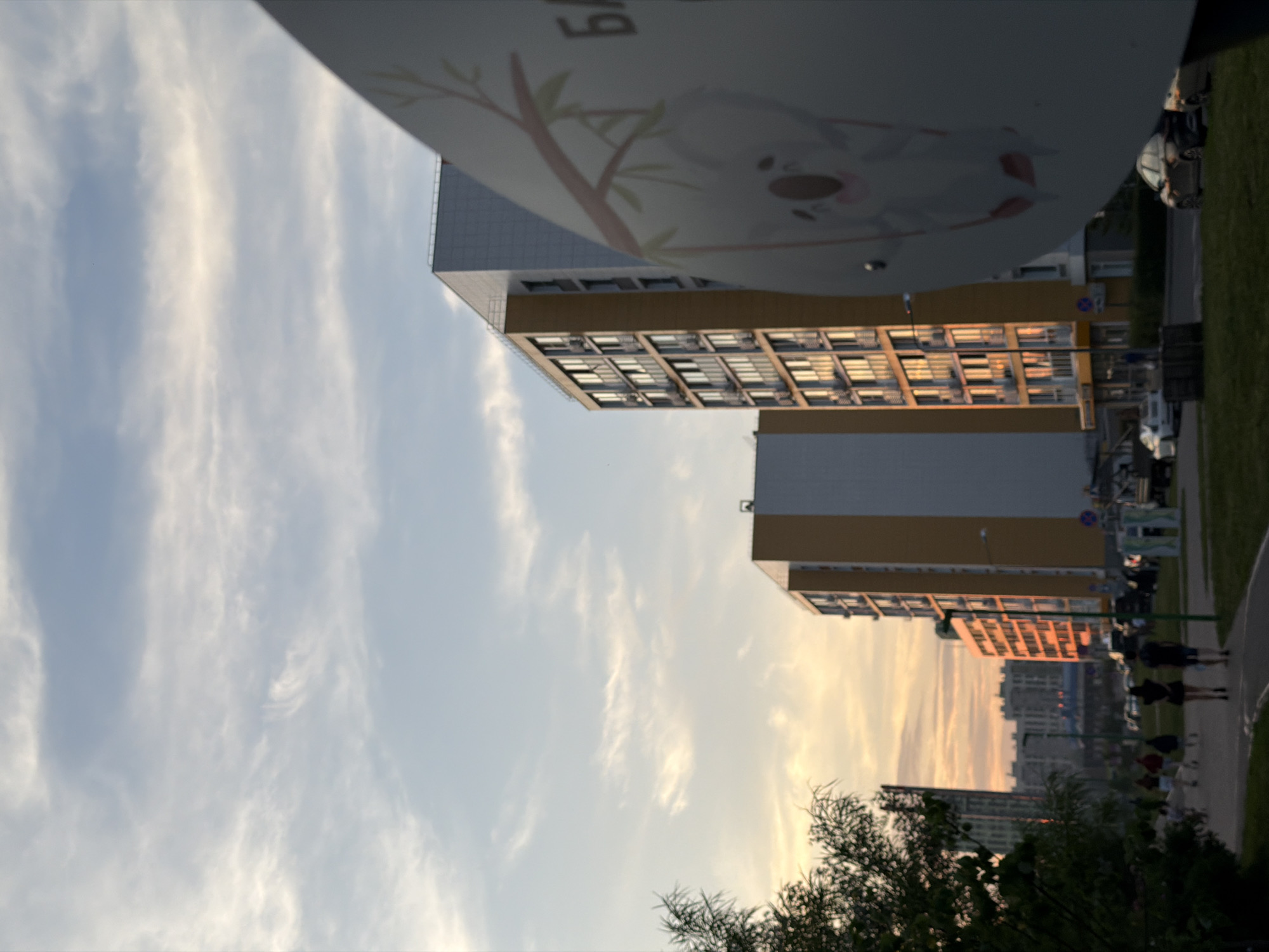 In an outdoor urban setting during sunset, a street scene unfolds with several modern apartment buildings as the primary backdrop. The sky displays a gradient of soft, wispy clouds transitioning from pale blue to warm orange hues near the horizon, indicating the sun is setting. In the foreground, a large, rounded object with a cartoon koala on it is prominently positioned, partially obscuring the view of the buildings. To the left, a cluster of trees with dark green foliage is visible, with some branches extending into the frame.
The apartment buildings are tall and contemporary, characterized by their colorful facades—some with yellow accents and others with white. Windows are visible on each floor, and some appear to have air conditioning units installed. The setting sun casts a warm, orange glow on the side of one of the yellow buildings, highlighting its architectural details.
On a paved walkway in front of the buildings, several people are walking, their figures silhouetted against the twilight. Two individuals, a man and a woman, are walking away from the viewer, with the man wearing shorts and a dark shirt and the woman in a dark dress. Other figures are further down the path, their details less distinct. Cars are parked along the street and on the road, with some moving. The ground is paved, with a grassy area in the immediate foreground where the large koala-themed object is situated. The overall impression is a tranquil evening scene in a residential urban area.