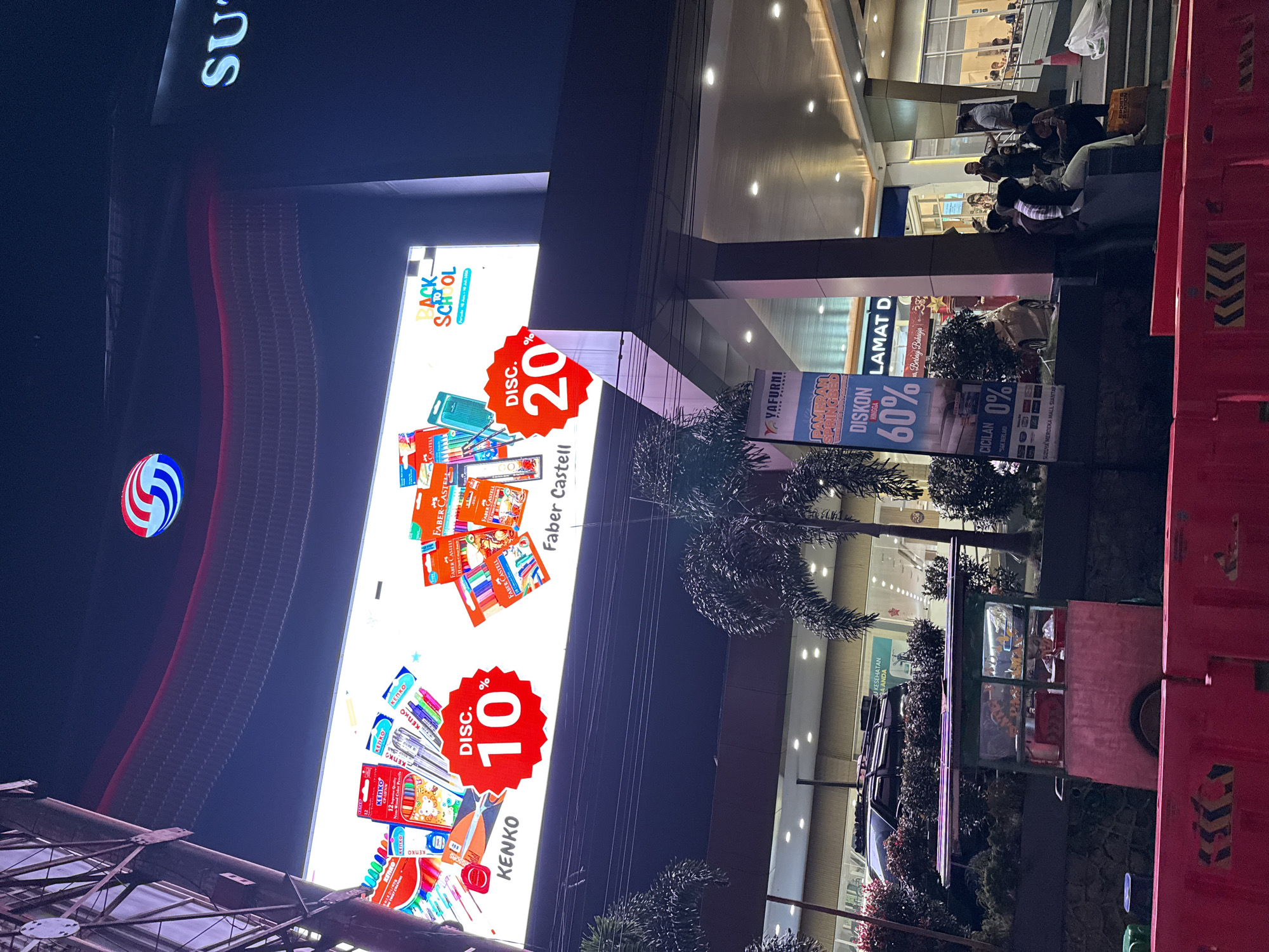 This is a vertical, nighttime photograph taken in Pematang Siantar, Indonesia, likely at a shopping mall or large retail complex. The scene is illuminated by bright LED displays and interior lighting.
Dominating the left side of the frame is a large, vibrant advertisement for a "Back to School" sale, featuring various art supplies like colored pencils and markers from brands such as Faber-Castell and Kenko. The advertisement prominently displays discounts of "DISC. 20%" and "DISC. 10%". Above this, a large, illuminated sign with the partial text "SU" is visible.
To the right, the entrance to a building is visible, with shoppers seated on steps outside. Further to the right, there are more advertisements. One banner announces "DISKON HINGGA 60%" (Discount up to 60%) and another promotes "CICILAN 0%" (0% Installments). The text "YAFURNI" is also visible on a sign. In the foreground, a food cart with displayed snacks is parked. The overall atmosphere appears to be lively and commercial, with people engaged in shopping or resting. The lighting suggests it is evening or night.