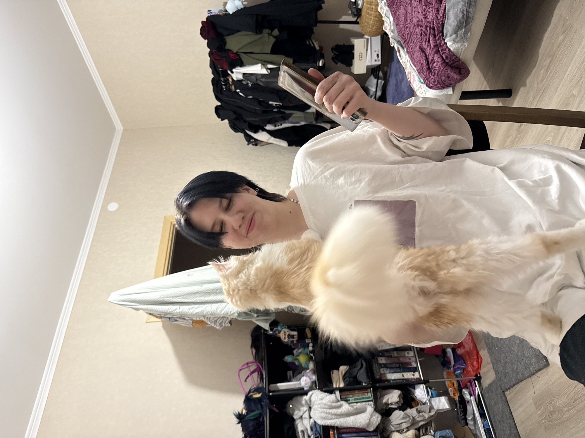 The image depicts an indoor domestic scene within a room located in Saint Petersburg, Russia. A young individual with short dark hair, wearing a white t-shirt, is seated and holding a cream-colored, long-haired cat. The person is looking towards the cat with a slight smile or smirk, their eyes partially closed. The cat is standing on its hind legs against the person's chest and left shoulder, looking upwards. The individual's right hand holds a rectangular silver-framed object, possibly a smartphone, vertically. A tribal-style tattoo is visible on the person's left forearm, and a silver chain is worn around the neck.
The room's environment features light beige textured wallpaper and a white ceiling with crown molding. The floor is light wood laminate. To the left, a black multi-shelf unit is cluttered with books, folded grey textiles, and various small objects, including a purple plastic item, a blue dragon figurine, and a white stuffed animal. Above the shelving unit, a striped towel or garment is draped over a light wooden door frame. To the right of the person, a significant pile or rack of dark-colored clothing, including black and red garments, is visible. Adjacent to this, a bed with a purple patterned blanket is partially shown, and a wicker basket, cardboard boxes (one with "Vans" branding), and a dark blue rug are on the floor.