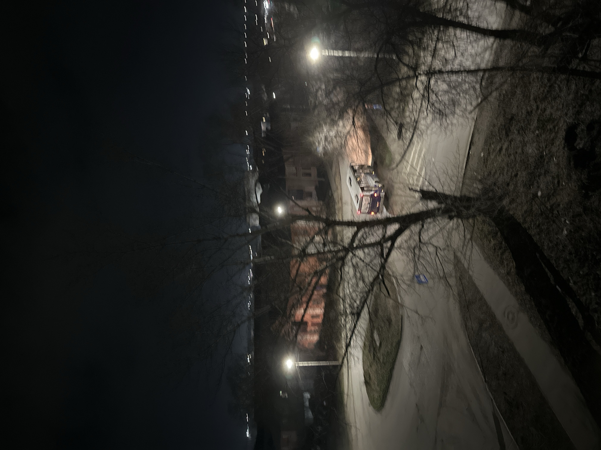 The image depicts a night scene in an urban environment. A street runs diagonally across the frame, illuminated by streetlights. Trees with bare branches frame the scene, obscuring the view partially. A vehicle, possibly an emergency response vehicle due to its coloring, is visible on the street. Lights from buildings are seen in the background, indicating a city or town setting. The location is in the city of Unknown, Unknown.