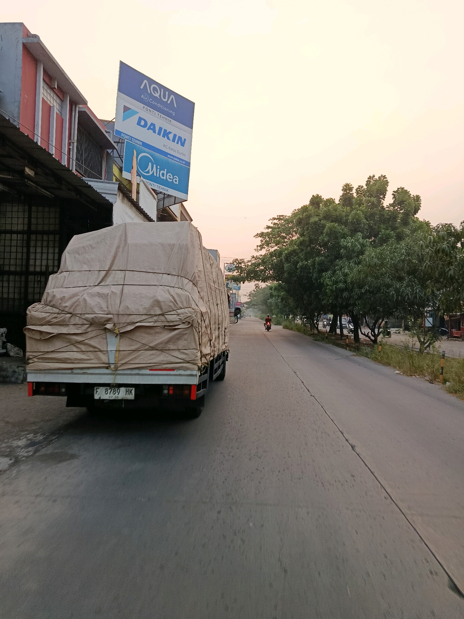 An outdoor, eye-level, medium shot captures a truck loaded with goods covered in a tan tarp on a paved road. The truck is moving away from the camera and is on the right side of the frame. The license plate reads "F 8789 HK". The truck is in an urban environment with a building on the left side of the frame featuring a large sign with logos for AQUA, DAIKIN, and Midea, indicating it is likely a retail or distribution center for these air conditioning brands. The building has red and grey facades and a dark metal security gate. Adjacent to the building is a black tarp.
On the right side of the road, a line of trees is visible, with some of them casting shadows on the road. Beyond the trees, there are other vehicles, including a motorcycle with a rider, parked cars, and possibly other shops. The sky is a pale orange and yellow, suggesting either sunrise or sunset. The road has a visible seam down the middle and appears somewhat worn. The presence of multiple air conditioning brand advertisements on the building strongly suggests this scene is located in an area known for commercial activity, potentially a hub for appliance sales or services. The street address or specific location within Bekasi, Indonesia, cannot be determined from the visual information alone, but the advertisements suggest a commercial district.