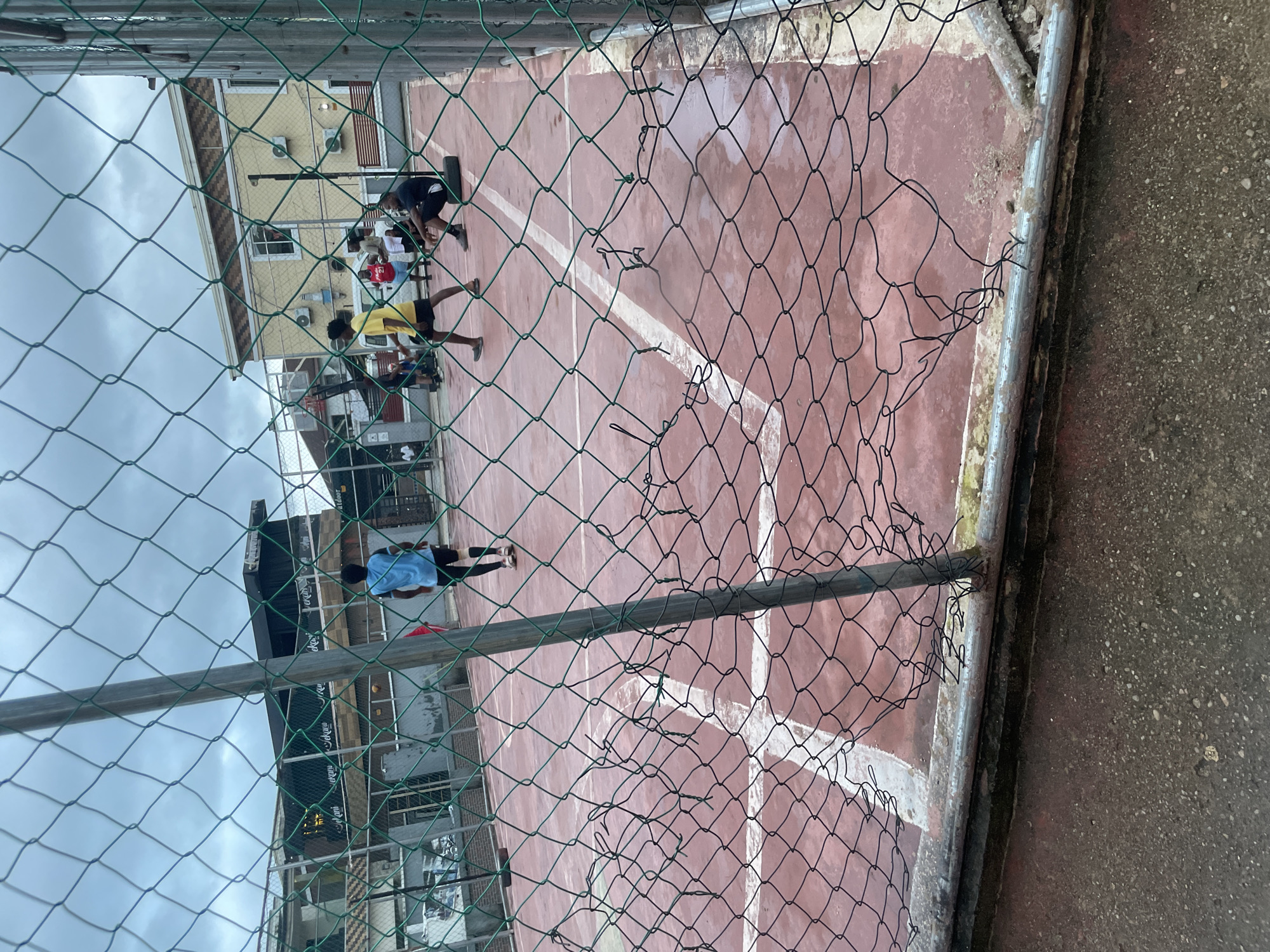 The image depicts a court with a reddish-brown surface and white markings, resembling a sports court, likely for basketball or volleyball. The court is enclosed by a green chain-link fence, parts of which are damaged and torn. In the background, there are buildings, including a yellow two-story structure with visible air conditioning units and windows, and another building with a darker facade and signage that reads "Sekani Sekani".
Several individuals are present on the court. In the center of the frame, a person wearing a blue jersey and black shorts is standing on the court. To their left, another person in a yellow t-shirt and black shorts is in motion, possibly playing. Further back, a group of people are scattered around the court. One individual is seated on a tire. The ground appears wet in places, suggesting recent rain. The sky is overcast and cloudy. The setting appears to be an outdoor recreational area within a residential or urban environment in Ihiagwa, Nigeria.