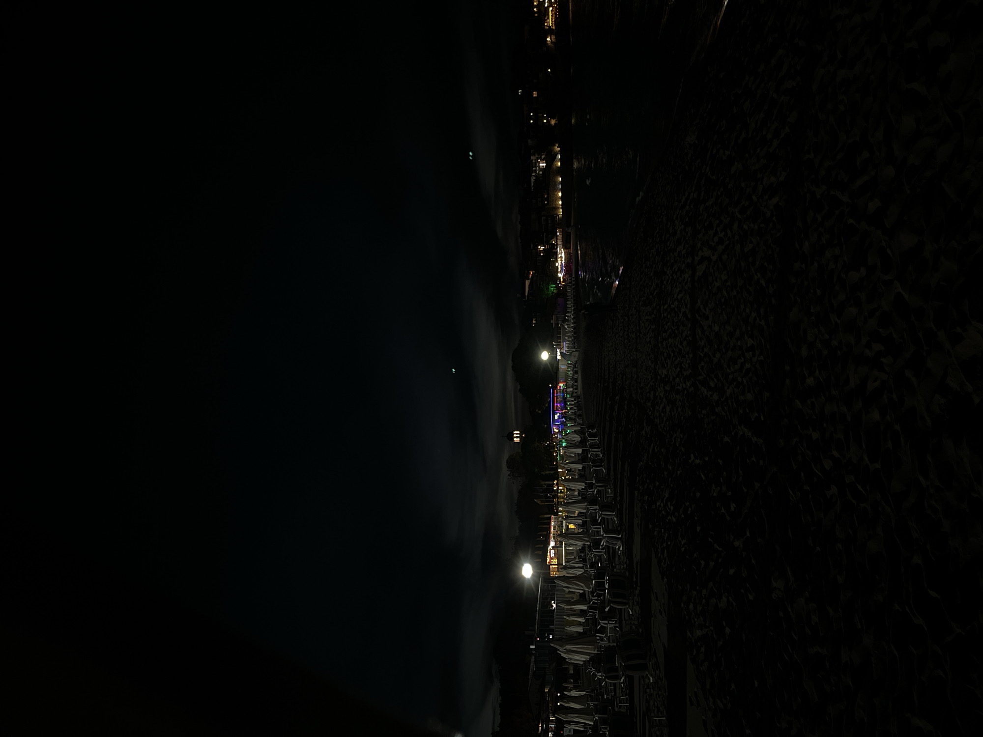 The image depicts a beach at night in Sozopol, Bulgaria. In the foreground, rows of closed beach umbrellas and chairs are set up on the sand. Further back, illuminated buildings with colorful lights line the waterfront. The sea is visible on the right, reflecting the distant lights. The sky is dark with some cloudy formations and faint lights, possibly distant lights or celestial bodies. The overall scene suggests a quiet, closed-down beach resort area after hours.
