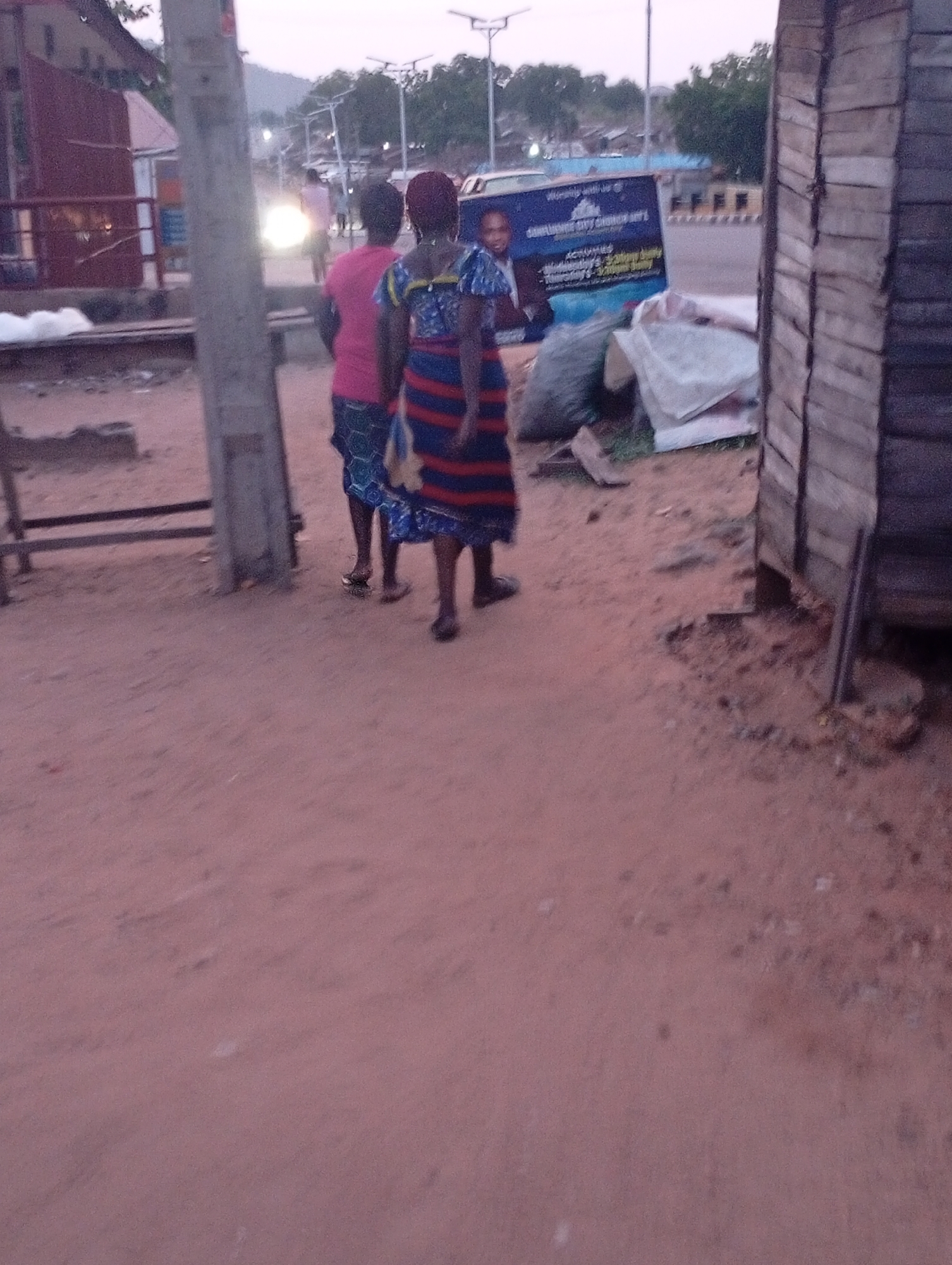 Two women walk away from the camera on a reddish-brown dirt surface in Zango-ndaji, Nigeria. The woman closer to the camera wears a patterned blue and red dress, while the other wears a pink shirt. In the background, there is a vehicle with an advertisement for Sumunte City Church, bags of garbage, and a wooden structure. There are streetlights and distant buildings visible in the background. A utility pole is visible to the left.
