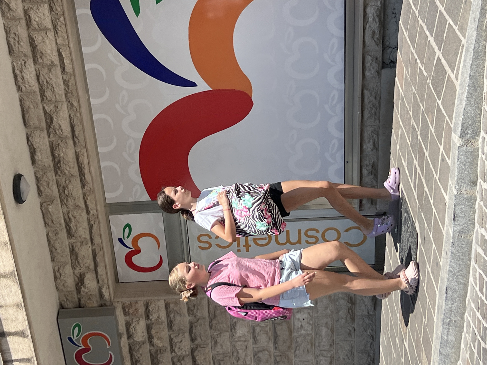 Two young girls are walking past a storefront in Budva, Montenegro. The girl on the left, with blonde hair pulled back in a bun, wears a pink t-shirt, denim shorts, and pink Crocs. She carries a pink backpack. The girl on the right, with brown hair, wears a white t-shirt, black shorts, and purple Crocs. She carries a large black and white patterned tote bag. The storefront features a large graphic with colorful abstract shapes and repeating apple logos. The building's facade is constructed of stone bricks. The girls are walking on a paved sidewalk.