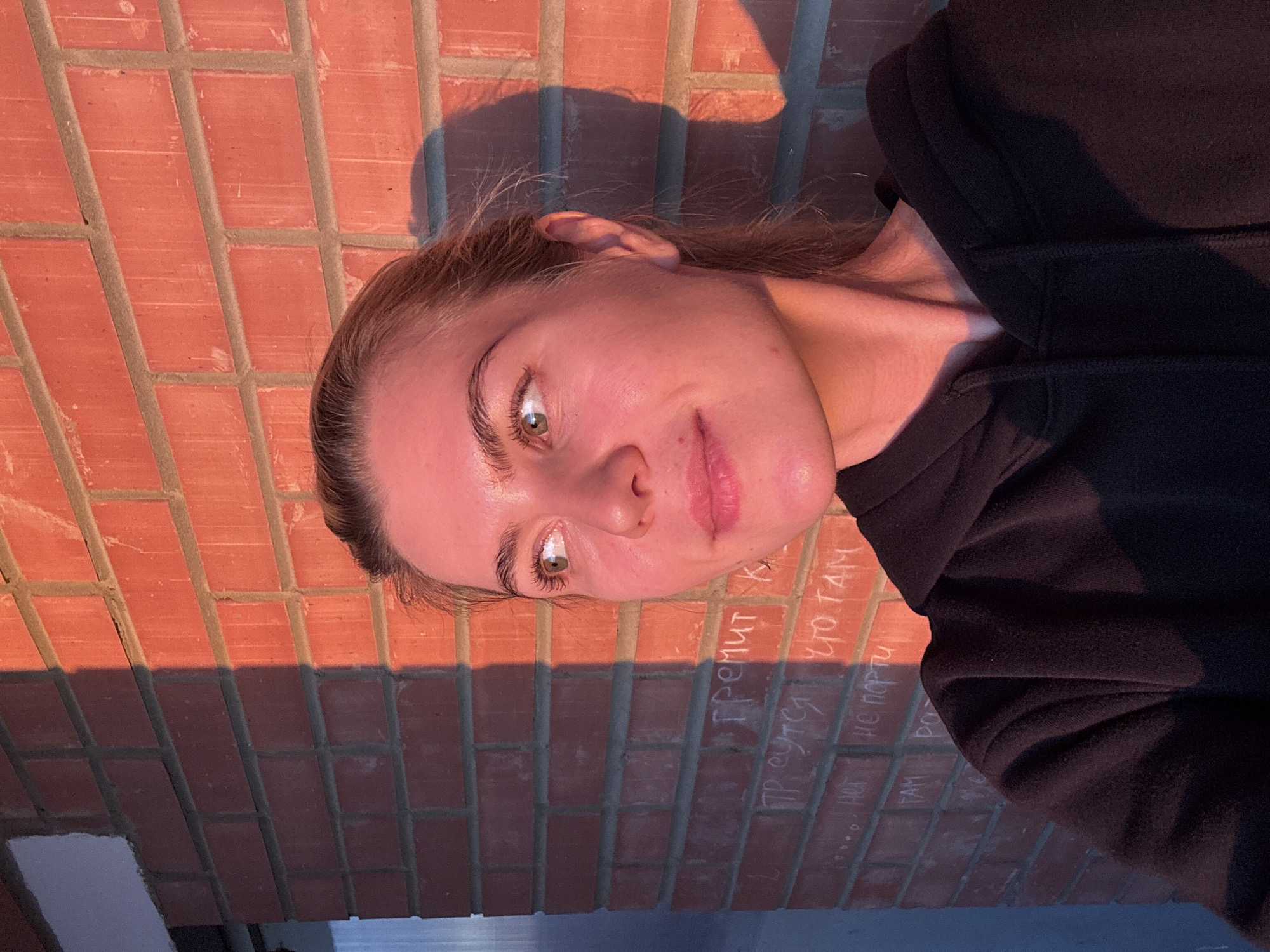 An outdoor, portrait-oriented shot captures a young adult female positioned to the left of the frame, illuminated by intense, warm-toned sunlight originating from the right. Her light-toned skin, particularly on the right side of her face and neck, reflects the golden-red hue of the light, contrasting with the cooler, shadowed areas on her left side. She has light-colored hair, partially pulled back, and green eyes, looking towards the upper right. Her lips are slightly upturned. She is wearing a black hooded sweatshirt.
The background consists of a red-orange brick wall with gray mortar lines. A sharp vertical line of shadow divides the wall, with the right side brightly lit by the warm light and the left side largely in deep, cool-toned shadow. Within the shadowed area of the brick wall, faint white Cyrillic graffiti is visible, including words such as "гремит" (thunders), "пр сутс" (present/s), "что там" (what's there), and "не порти" (don't spoil). The subject's shadow is cast distinctly on the illuminated portion of the brick wall to her right. To the far left, a shadowed, vertical structure, possibly a doorframe or an adjacent wall, is partially visible, appearing dark blue-gray. The location is Unknown, Unknown.