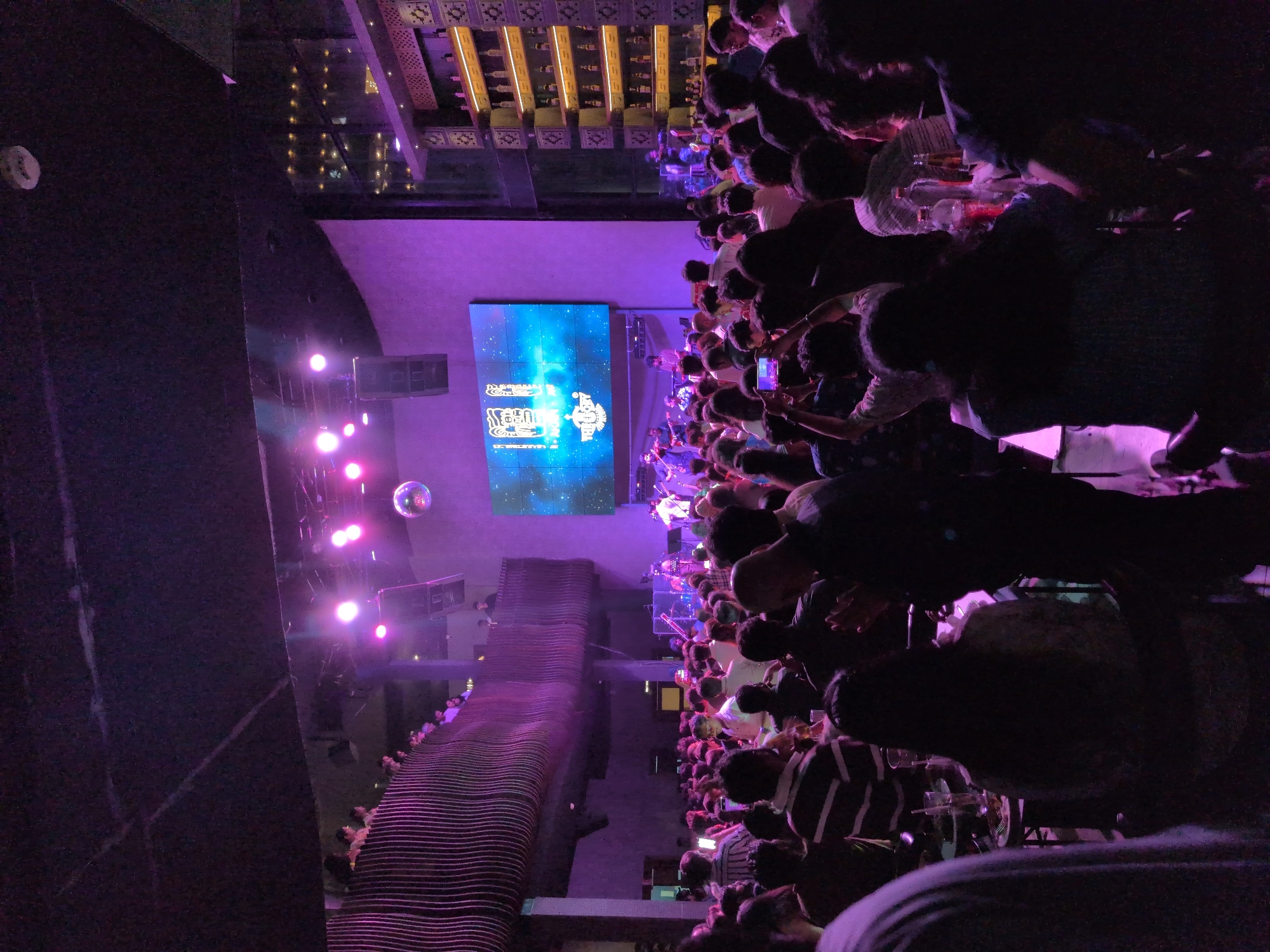 This detailed image depicts a crowded, multi-level indoor entertainment venue, likely a nightclub or bar, located in Hyderabad, India. The scene is dominated by vibrant purple and pink lighting, creating a dynamic nightlife atmosphere.
In the foreground and midground, a large assembly of approximately 100 to 150 individuals, predominantly young to middle-aged adults, occupies the main floor. Many are standing, observing a live musical performance. Several individuals are visible holding smartphones aloft, indicating they are recording or photographing the event. Scattered tables with beverages and residual food items are interspersed within the crowd.
At the rear of the venue, a raised stage hosts a live band composed of multiple musicians, actively performing with instruments including guitars, drums, and a keyboard. Behind the performers, a large LED screen displays stylized white graphics and the text "THE CITY" against a blue, cosmic-themed background. Large black speaker cabinets are mounted on either side of the stage.
An upper-level balcony extends along the left side of the venue, featuring a distinctive, wavy, vertical slatted railing structure, also illuminated with purple light. A smaller group of people is present on this elevated level, overlooking the main floor.
To the right of the main area, a prominent, multi-tiered shelving unit is integrated into the wall, filled with numerous bottles, presumably alcoholic beverages, and is illuminated from within. A spherical disco ball hangs from the dark ceiling, reflecting ambient light. The venue's walls exhibit a textured finish, appearing light purple under the pervasive lighting. The overall environment suggests a lively social gathering centered on a live musical performance.