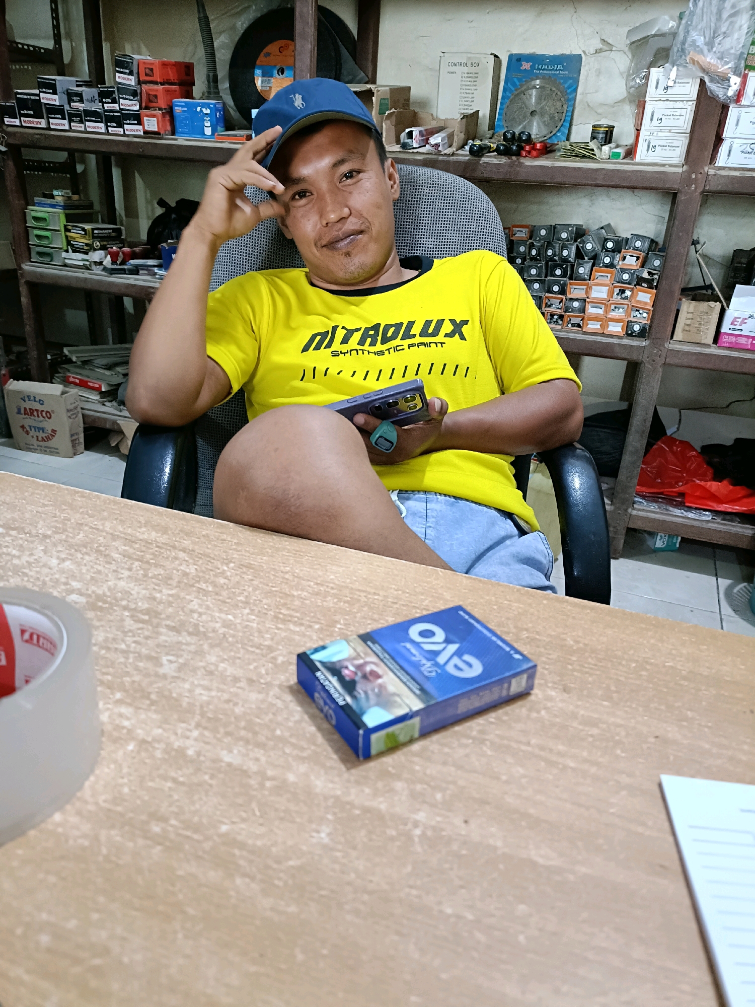 A medium shot captures a man sitting in a chair in what appears to be a retail or storage environment. He is wearing a yellow t-shirt with black text and a blue baseball cap. The man is holding a smartphone in his right hand and has his left hand raised to his forehead. He is looking towards the camera with a slight smile. In the foreground, on a wooden table, are a roll of tape, a pack of cigarettes, and a piece of paper. Shelves filled with various items, including boxes and what appear to be tools or hardware, are visible in the background. The scene suggests a casual setting, possibly a shop or workshop in Pordapor, Indonesia.