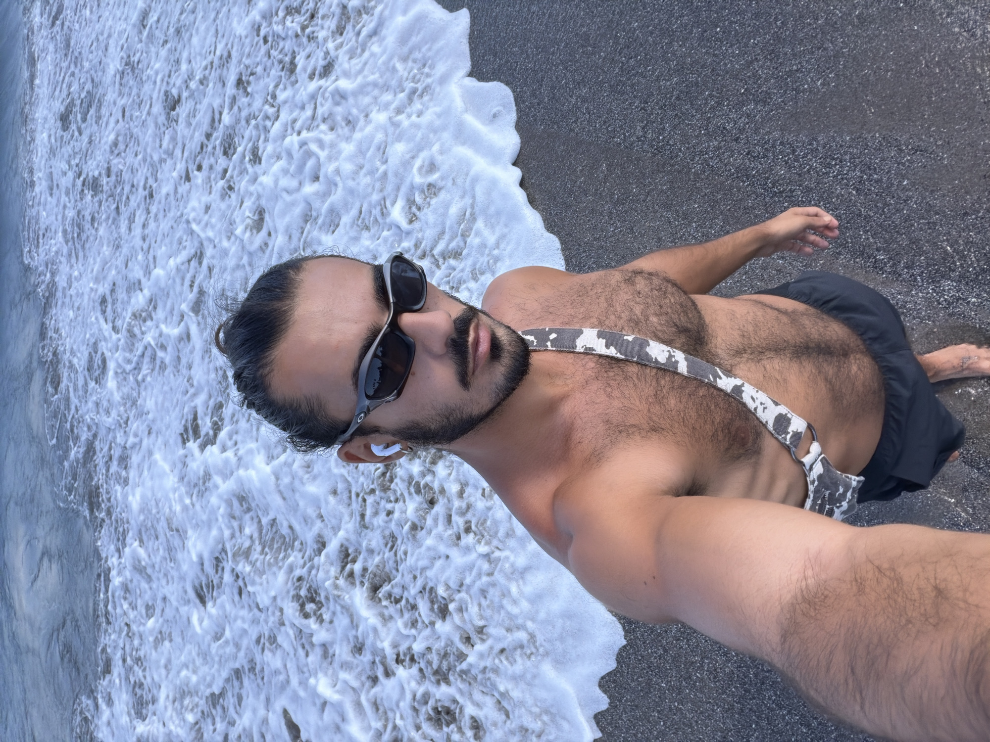 A man is standing on a dark sand beach with waves lapping at his feet in North Kuta, Indonesia. He is shirtless and wearing black swim shorts and a strap across his chest with a gray and white cow print pattern. He has sunglasses on and white earbuds in his ears, and his hair is pulled back in a bun. The water is foamy and white, with the ocean stretching out behind him.