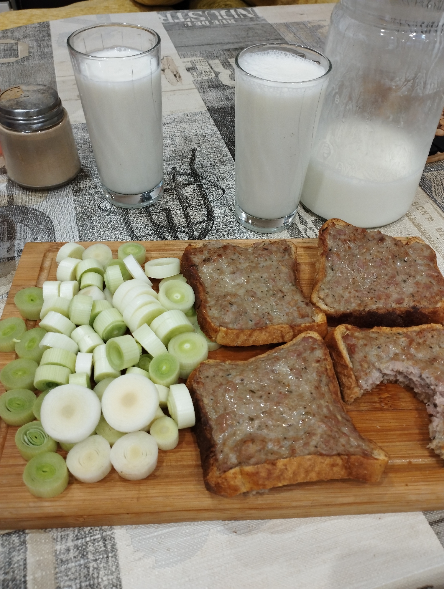 On a wooden cutting board on a table with a patterned tablecloth are sliced leeks and four pieces of bread topped with what appears to be sausage meat. Beside the board are two glasses filled with a white liquid, possibly milk, and a jar of the same liquid. A small salt shaker is near one of the glasses. The environment suggests a home setting. The location is Unknown, Unknown.