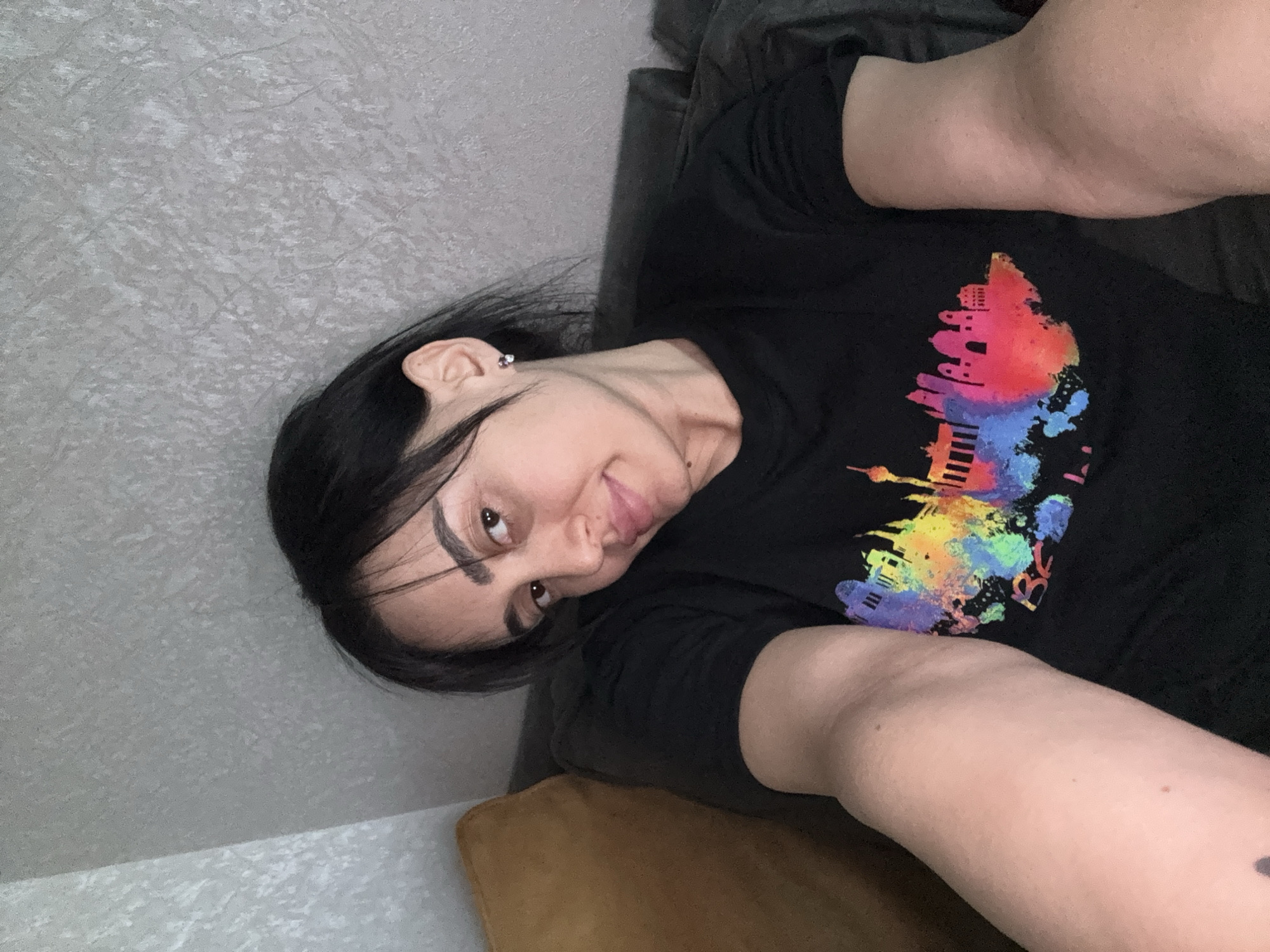 A woman is reclining on a couch. She is fair-skinned with dark hair and is wearing a black t-shirt printed with a skyline design in a variety of bright colors. The skyline appears to resemble the architecture of the city of Tbilisi, Georgia. The background includes a textured wall and a dark-colored couch.